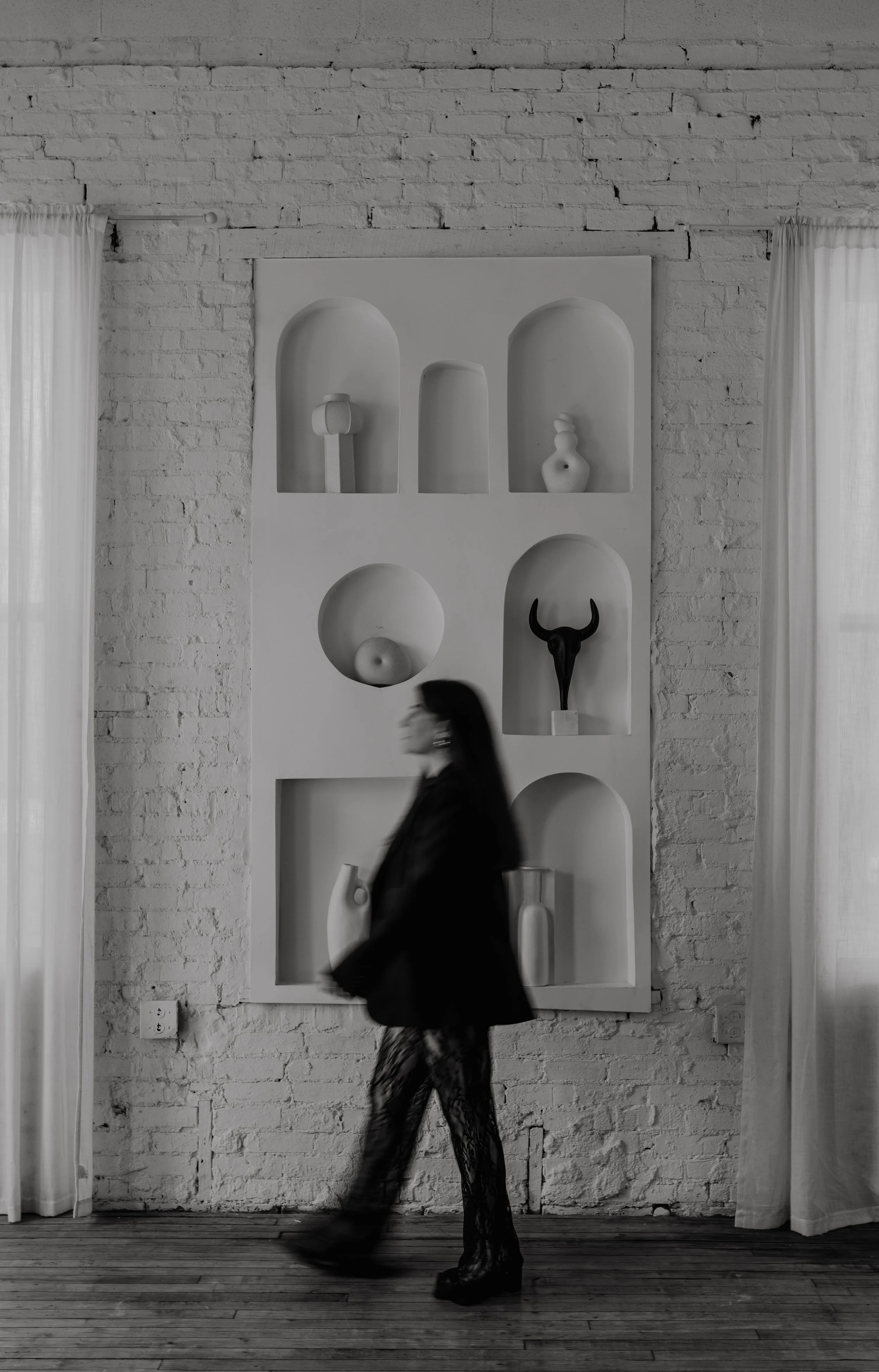 A woman walking past a white brick wall with a display shelf featuring minimalist sculptures and vases, in a modern art gallery setting.