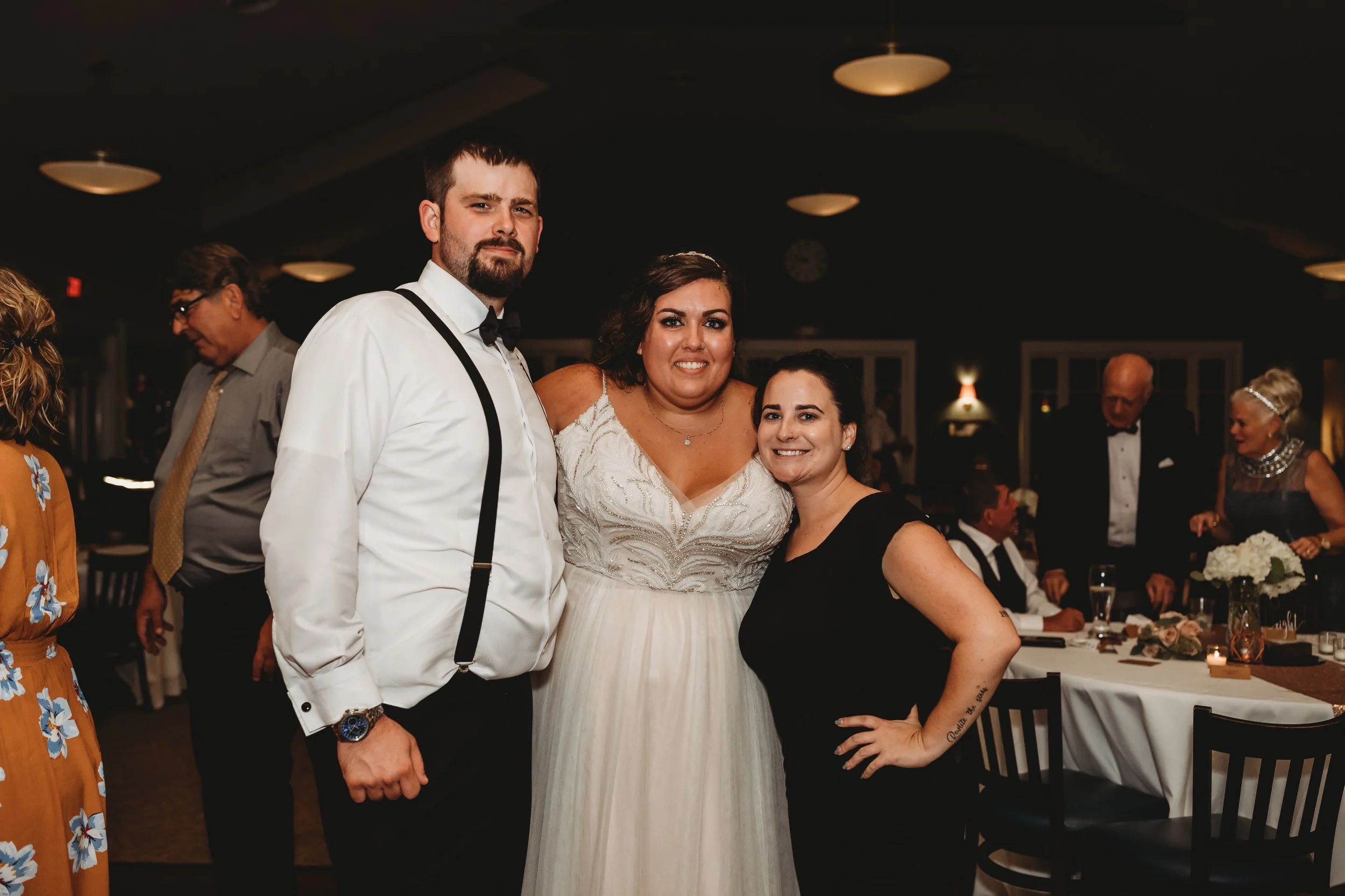 Group of three people, a man in a tuxedo, a woman in a white wedding dress, and another woman in black, smiling at a wedding reception.