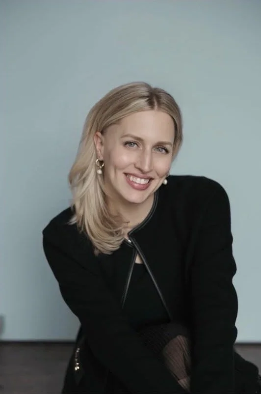 A woman with blonde hair smiling, wearing a black jacket and earrings, sitting against a light blue background.