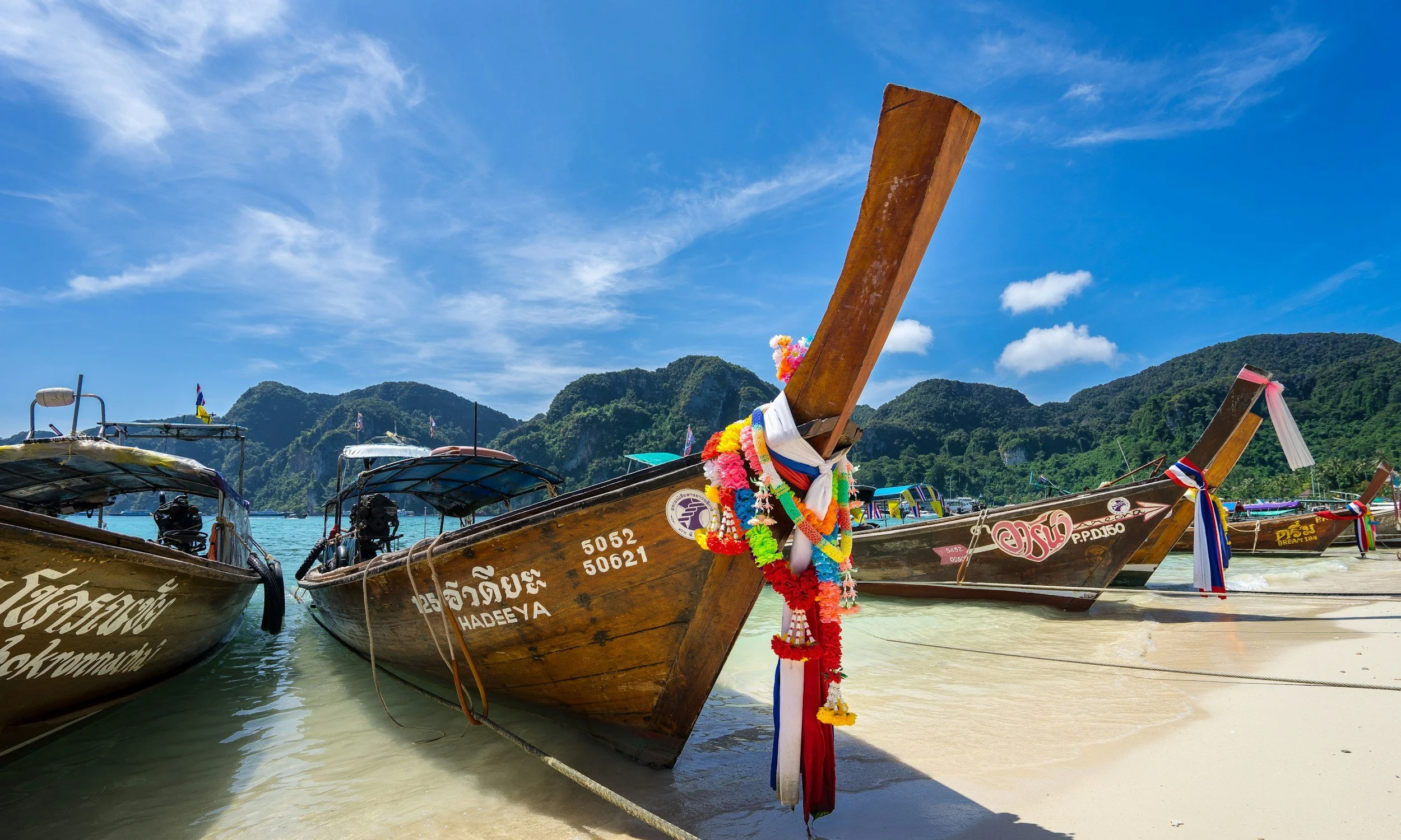 Bateaux en bois décorés avec des guirlandes colorées sur une plage avec des montagnes en arrière-plan, sous un ciel bleu avec quelques nuages.