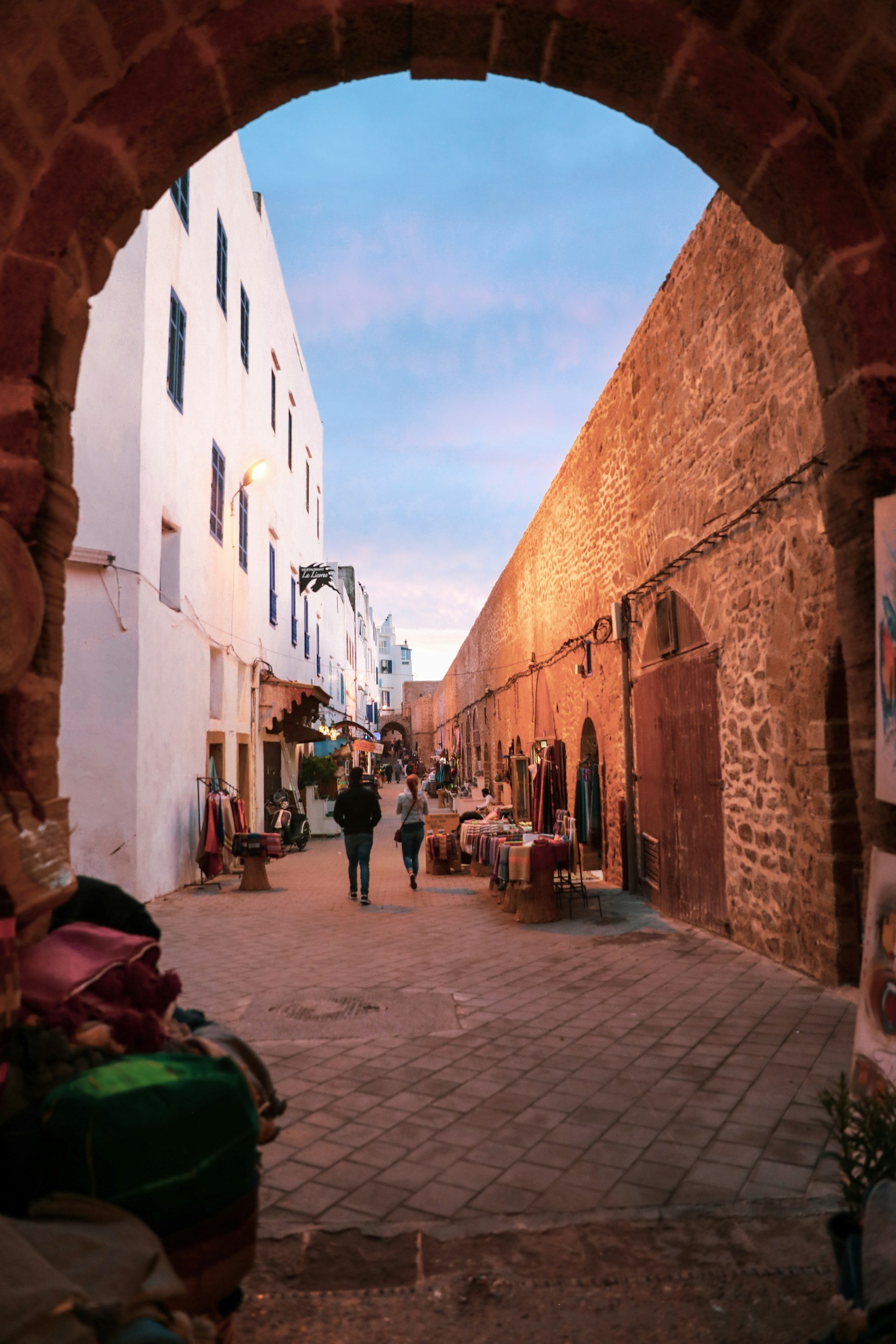 Une rue piétonne bordée de boutiques, vue à travers une archway en pierre, avec des murs blancs et en pierre, au coucher du soleil.