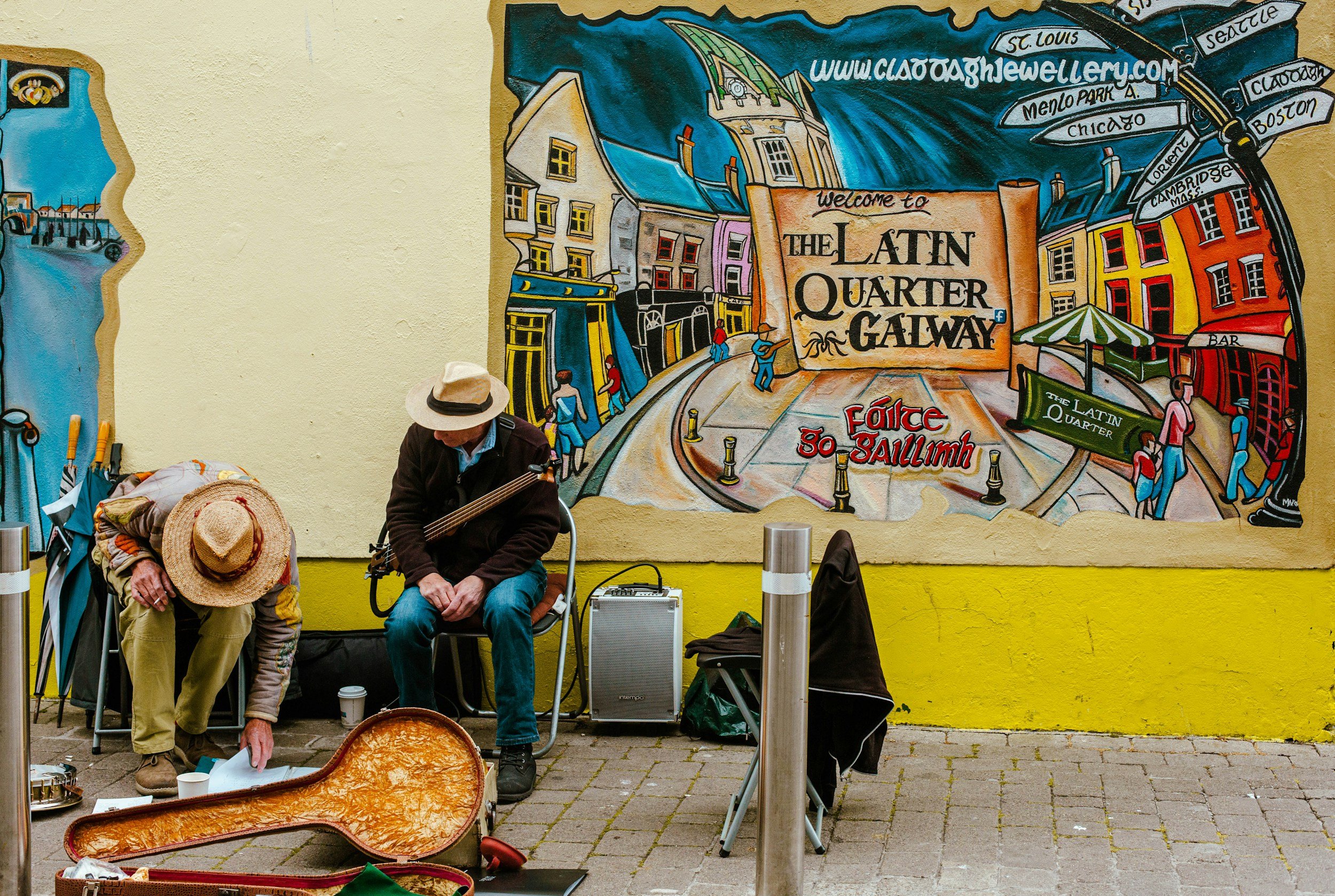 Deux musiciens de rue assis devant un mur peint représentant le quartier latin à Galway, en Irlande. Un homme est en train de jouer de la guitare et un autre organise ses instruments, avec une architecture colorée en arrière-plan.