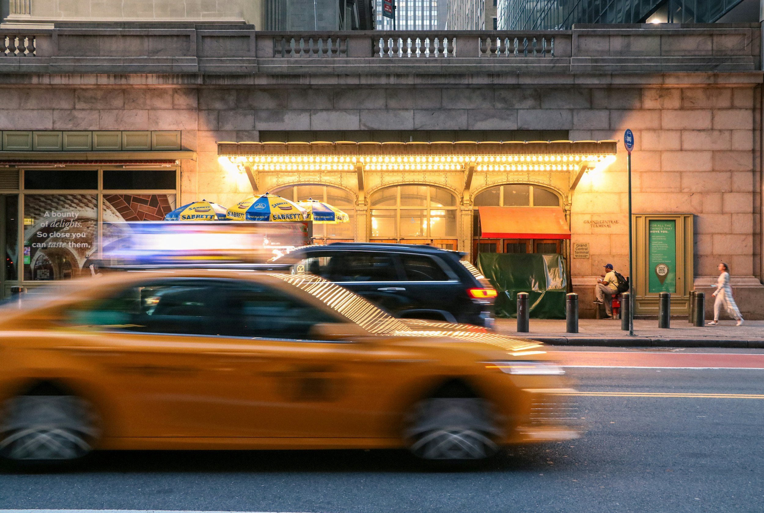 Vue urbaine avec des voitures en mouvement devant une façade de bâtiment éclairée, avec des parasols jaunes et bleus en terrasse et des personnes assises ou marchant le long du trottoir.