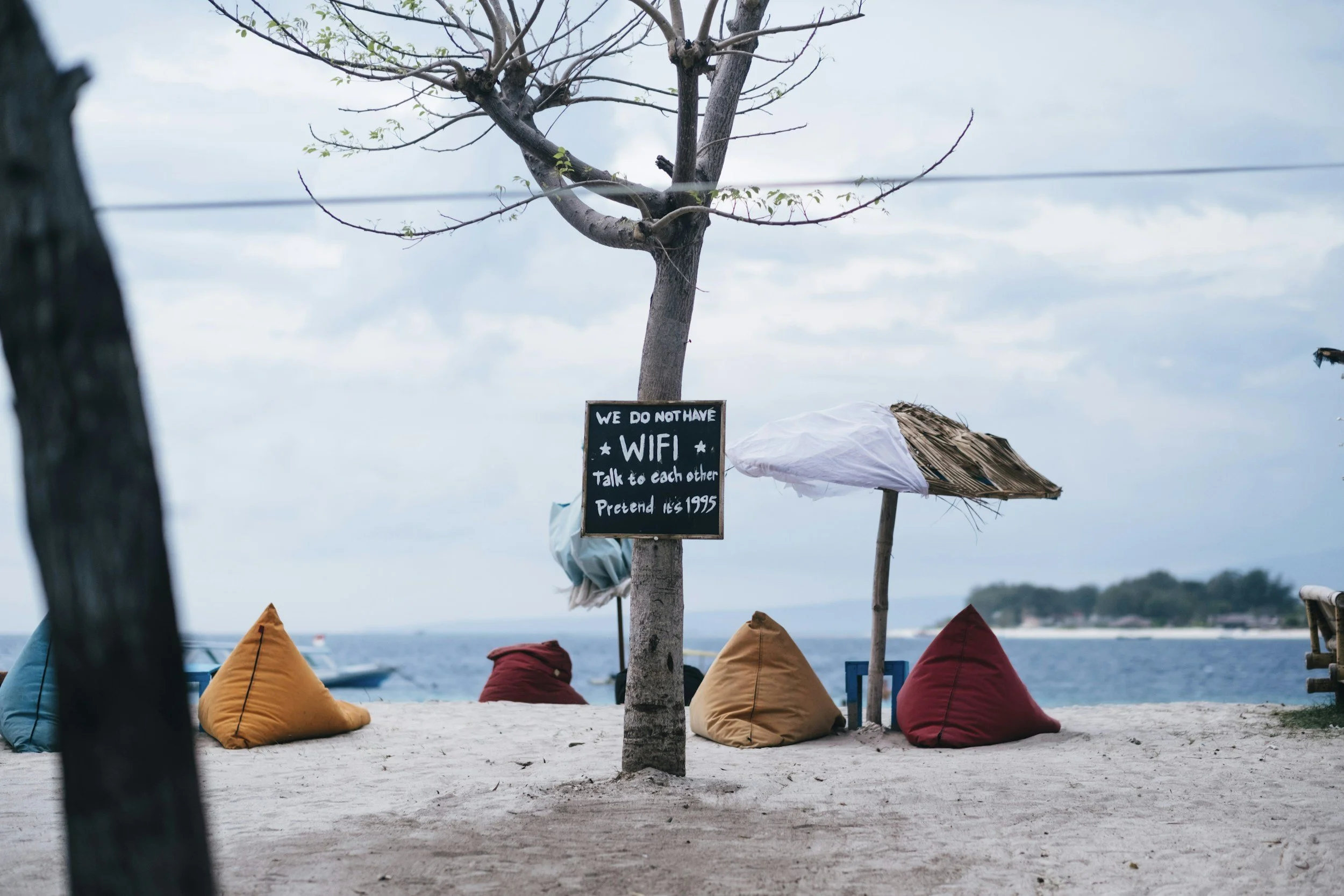 Une plage avec des coussins colorés sur le sable, un arbre avec un panneau indiquant 'We do not have WiFi', et des parasols en paille, avec la mer en arrière-plan.
