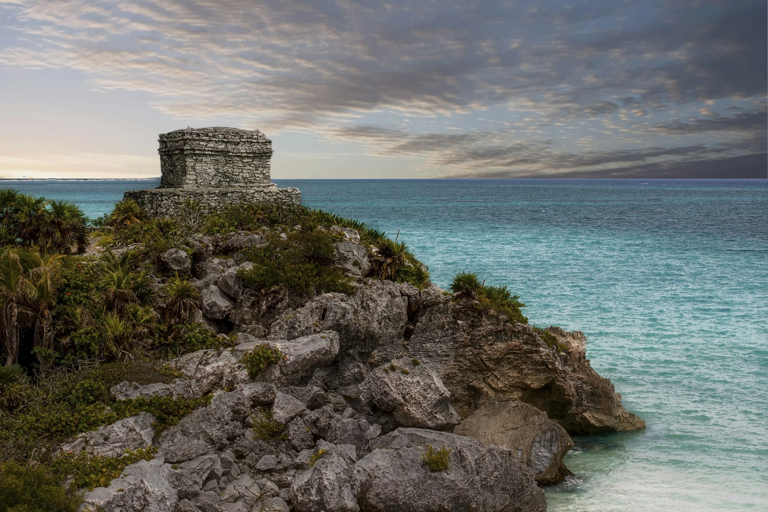 Tour en ruines de pierre sur une falaise rocheuse surplombant une mer turquoise, avec un ciel nuageux au coucher de soleil.