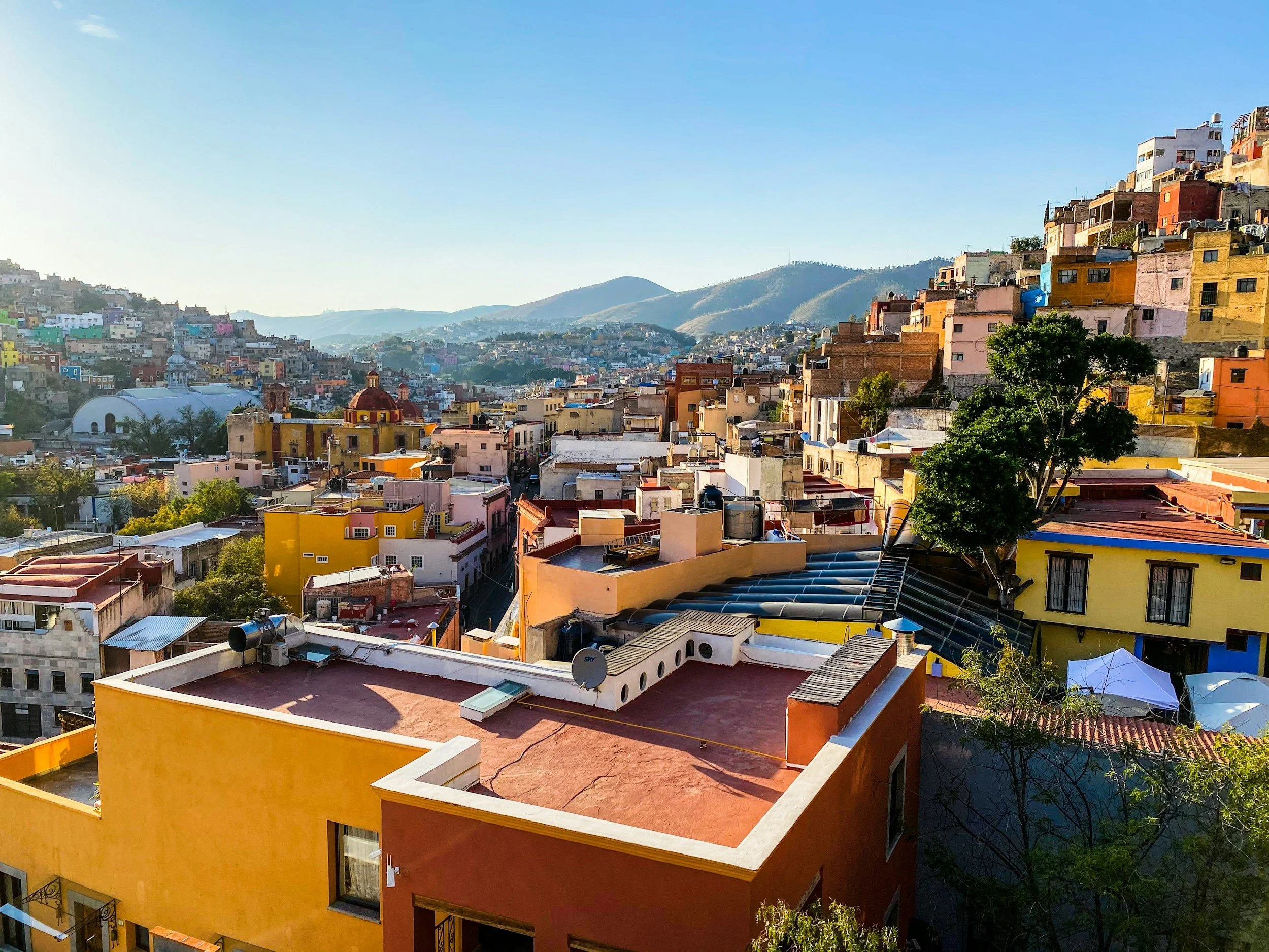 Vue panoramique colorée d'une ville construite en terrasses sur une colline avec des montagnes au loin, ciel clair et ensoleillé.