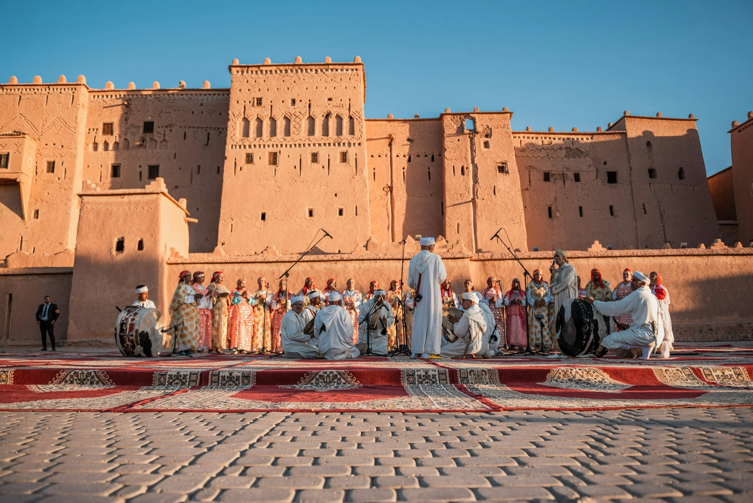Groupe de musiciens et chanteurs traditionnels marocains en train de performer devant un ancien kasbah en adobe dans le désert, avec des tapis colorés au sol. Lors du coucher de soleil.