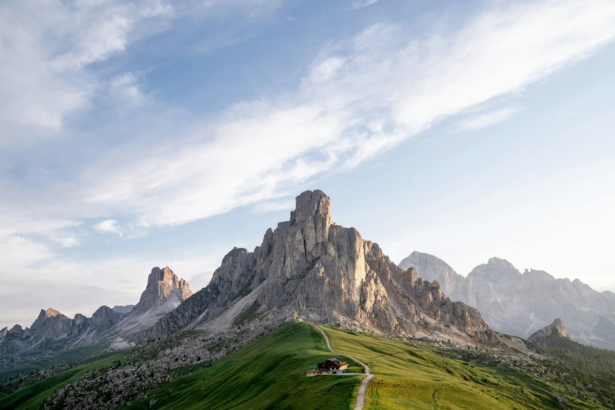 Paysage de montagnes avec sommet rocheux, prairies vertes en dessous et un chemin menant à une maison au centre