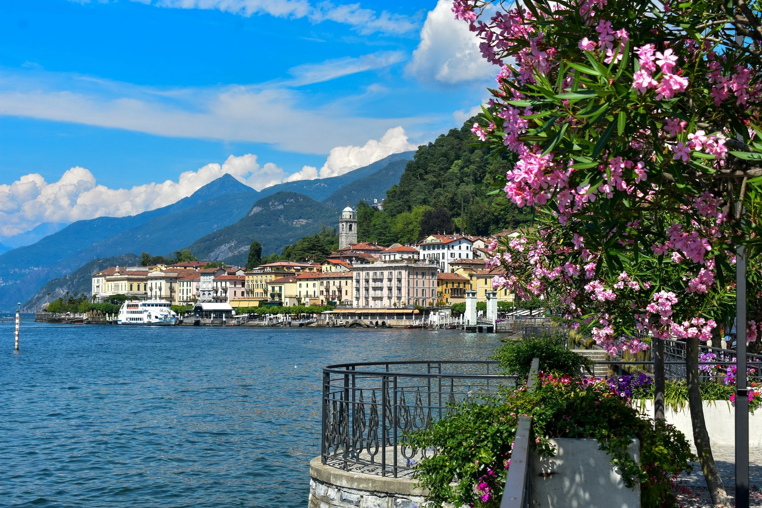 Vue d'un lac avec des bâtiments colorés, des montagnes en arrière-plan, des fleurs roses et une promenade au bord de l'eau.