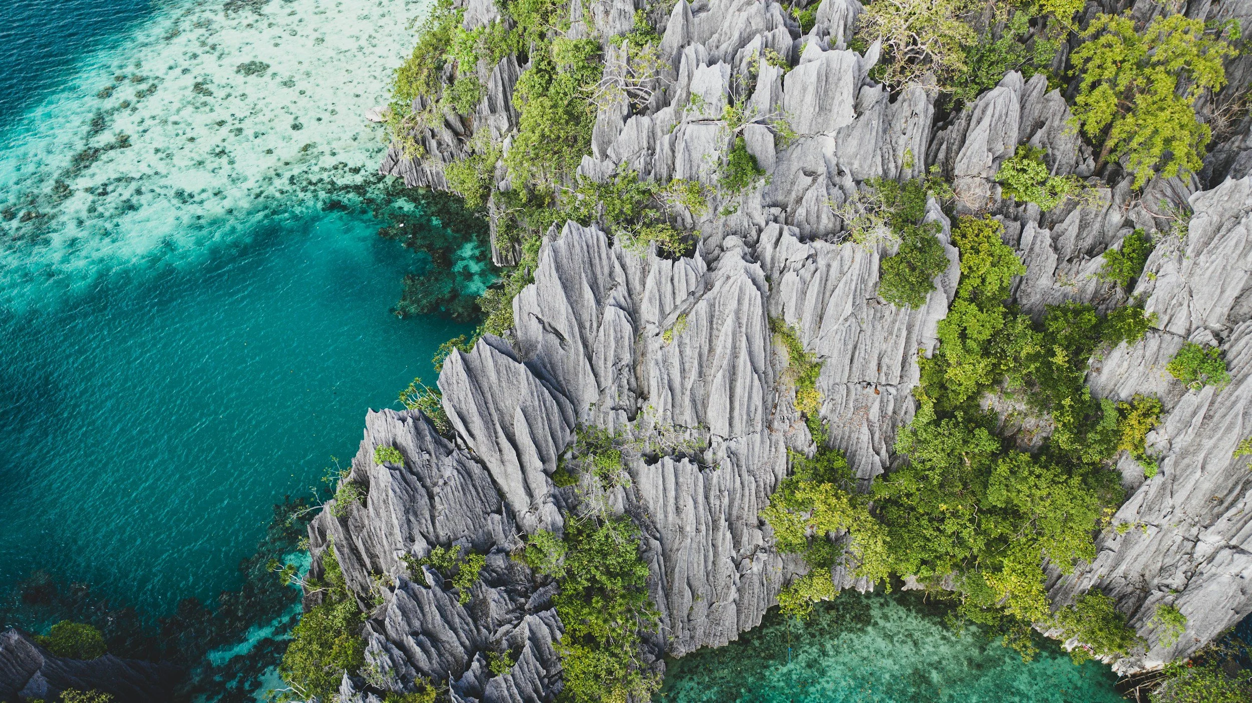 Photo aérienne de falaises rocheuses avec végétation verte surplombant une mer turquoise avec des eaux claires et un récif corallien visible par transparence.