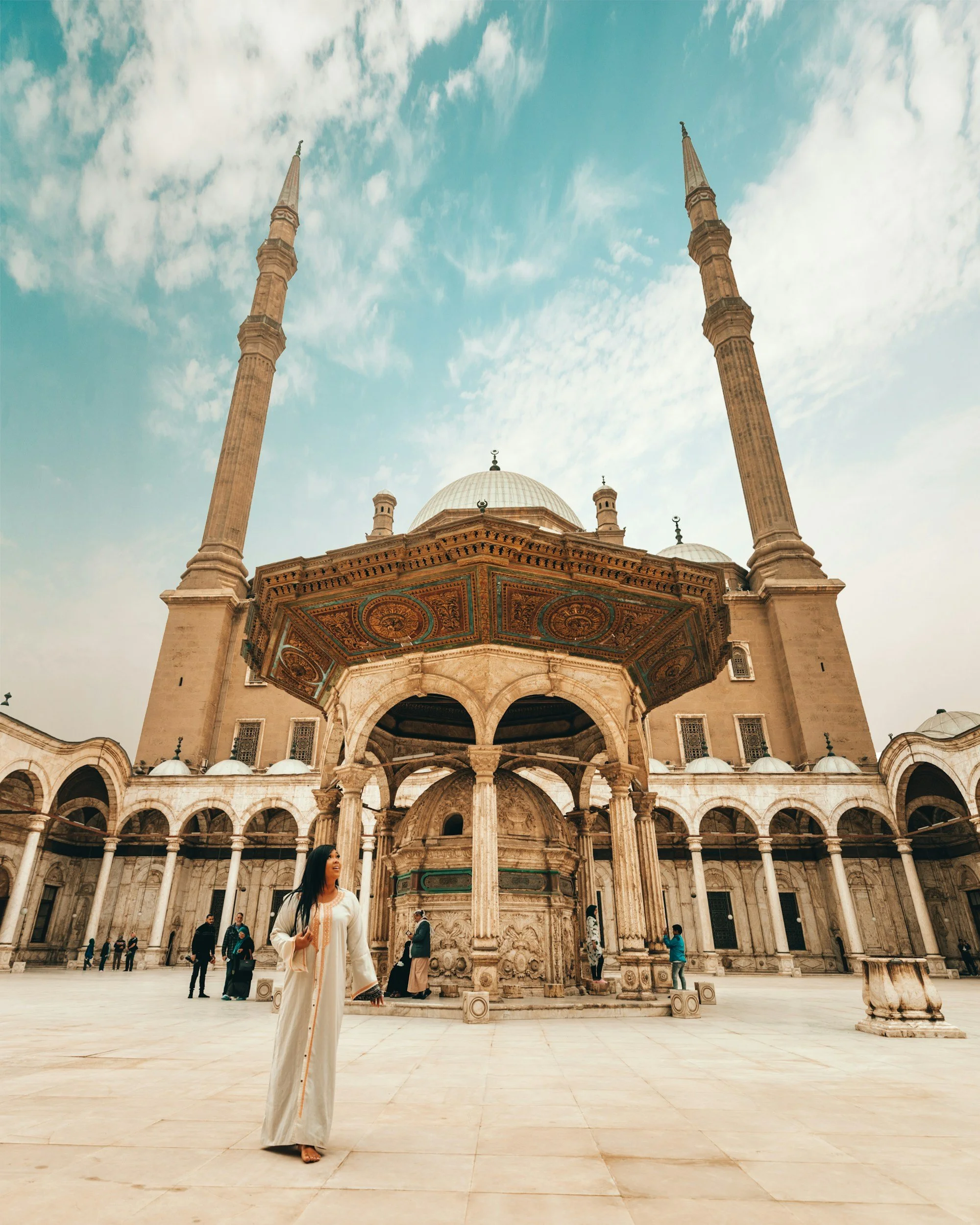 Une femme en robe blanche dans la cour d'une grande mosquée avec deux minarets, une coupole centrale, et une fontaine en pierre, sous un ciel partiellement nuageux.