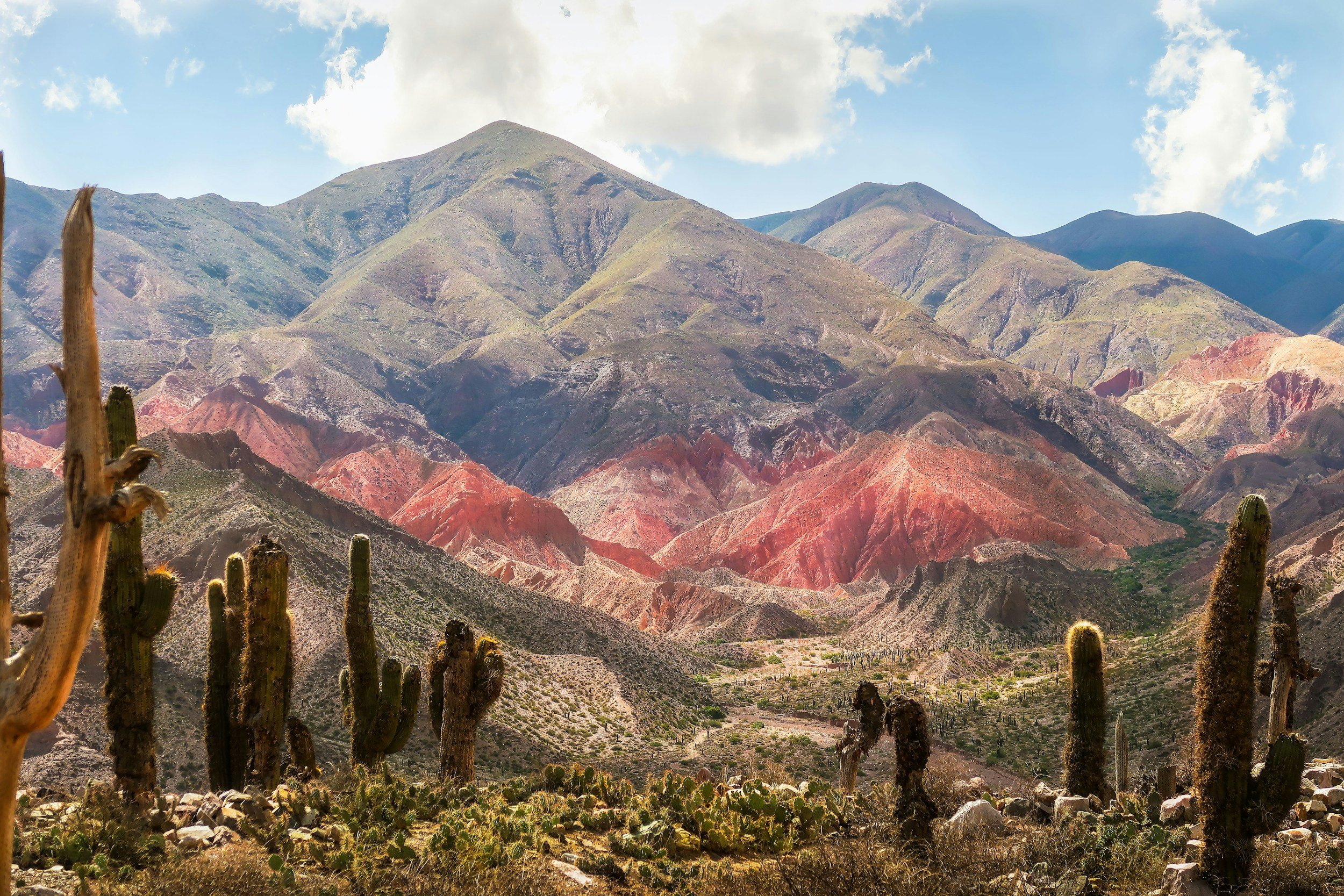 Paysage montagneux avec des sommets colorés, cactus au premier plan et ciel partiellement nuageux.