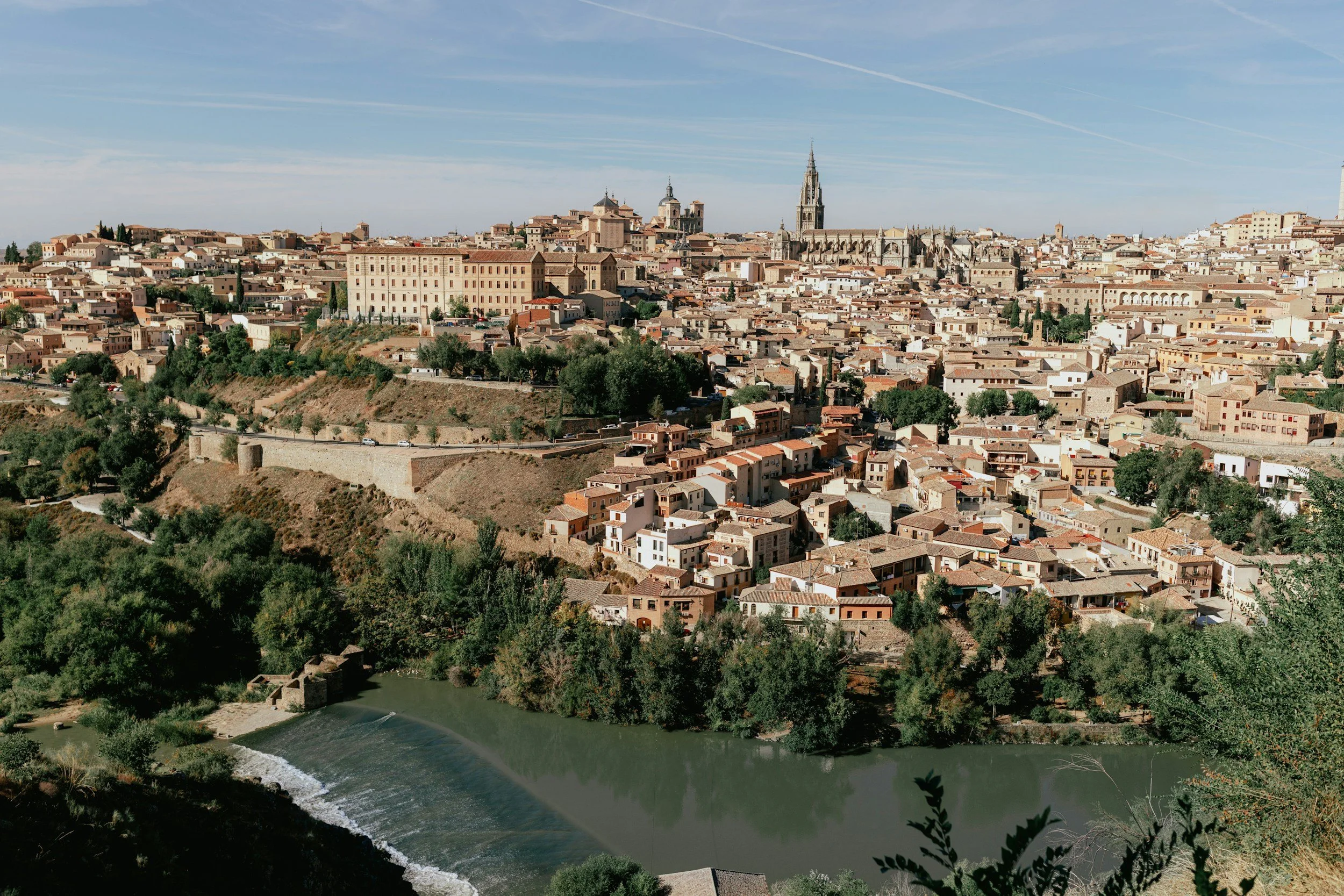 Vue panoramique de la vieille ville de Tolède avec ses bâtiments colorés, sa cathédrale gothique emblématique, et la rivière qui traverse la ville, sous un ciel clair.