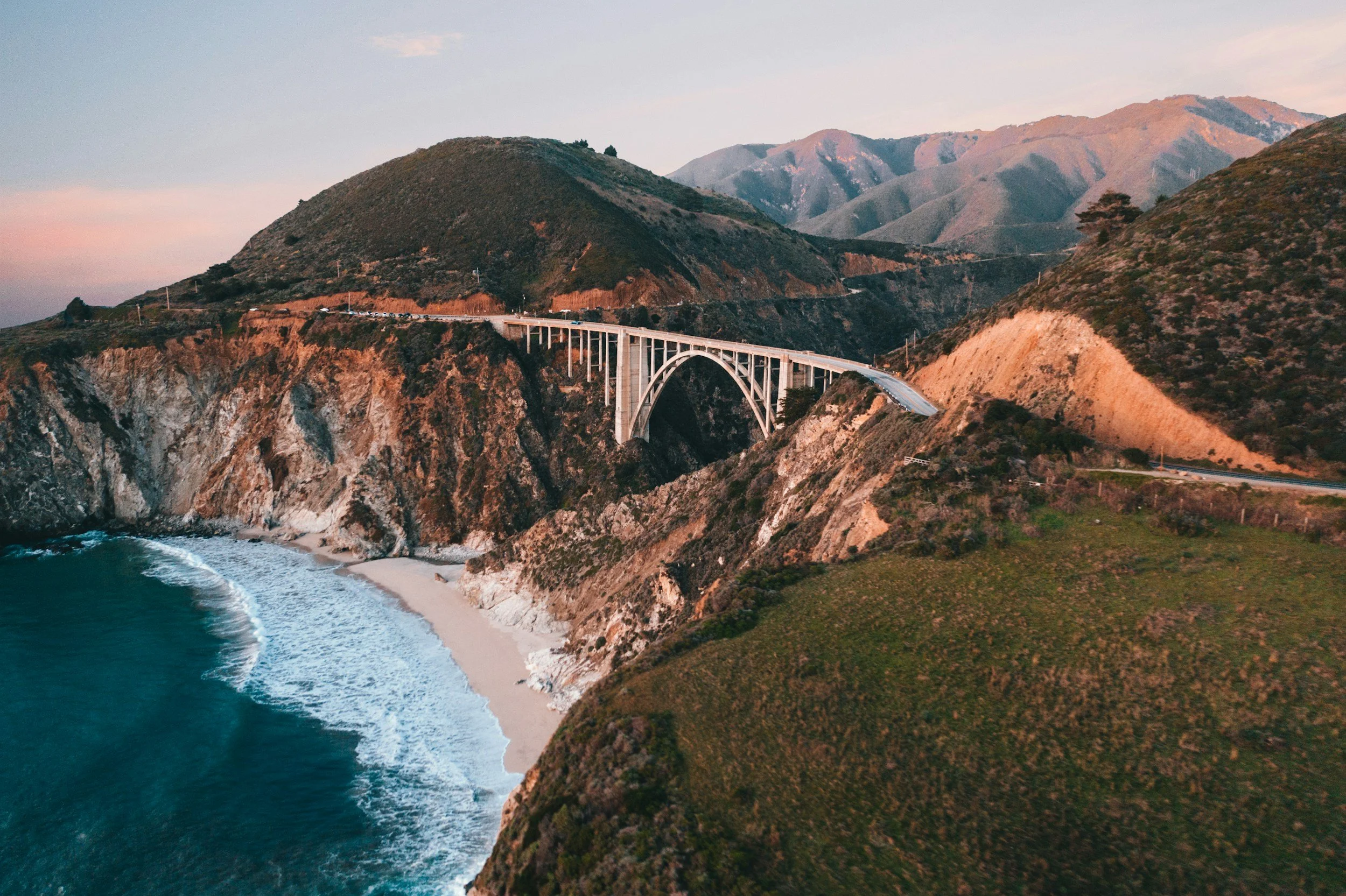 Un pont en arc construit en béton reliant deux falaises rocheuses au bord de l'océan, avec des montagnes en arrière-plan au coucher du soleil.