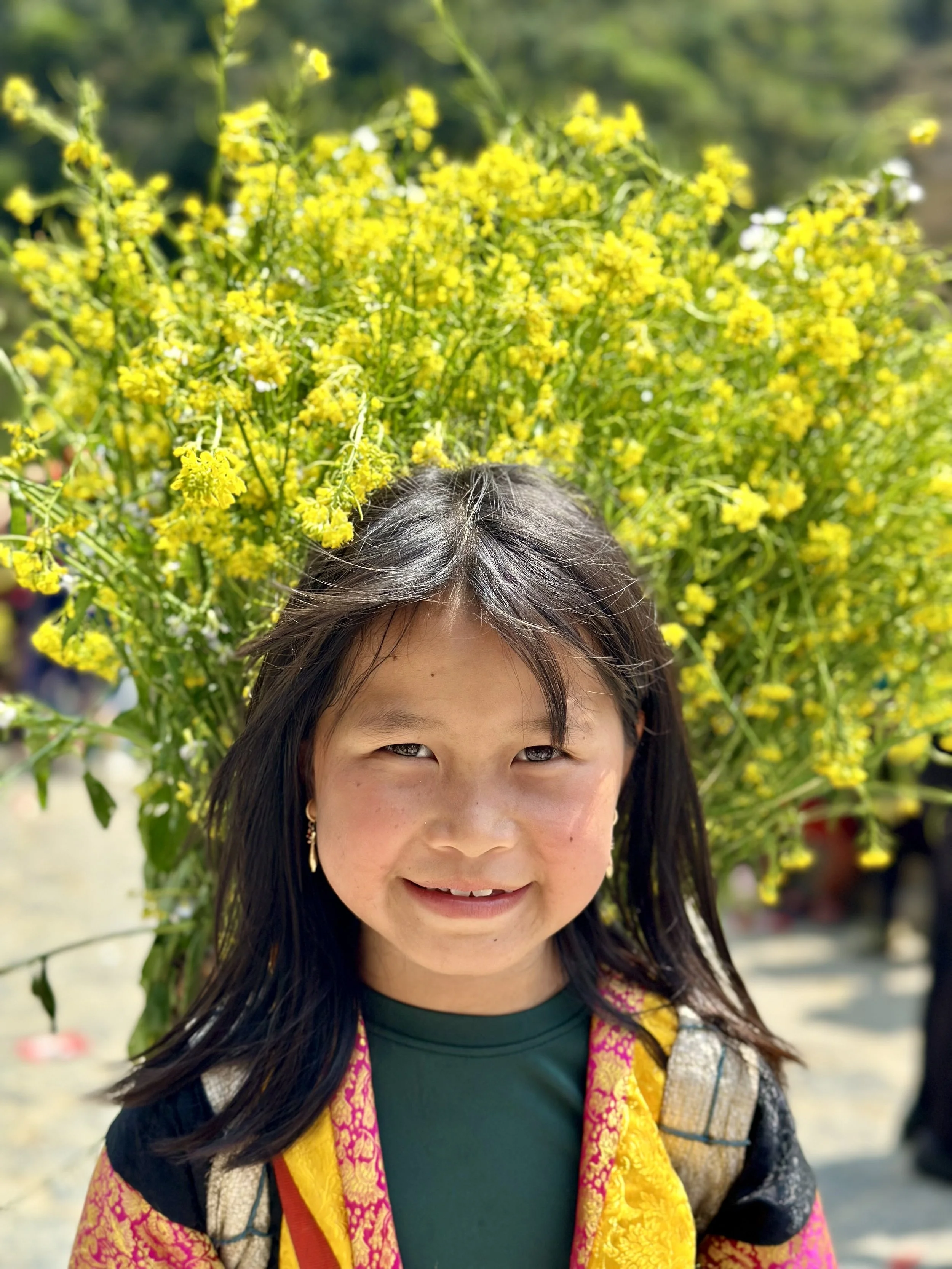 Jeune fille souriante avec un collier jaune, portant un sac à dos, devant un grand bouquet de fleurs jaunes.