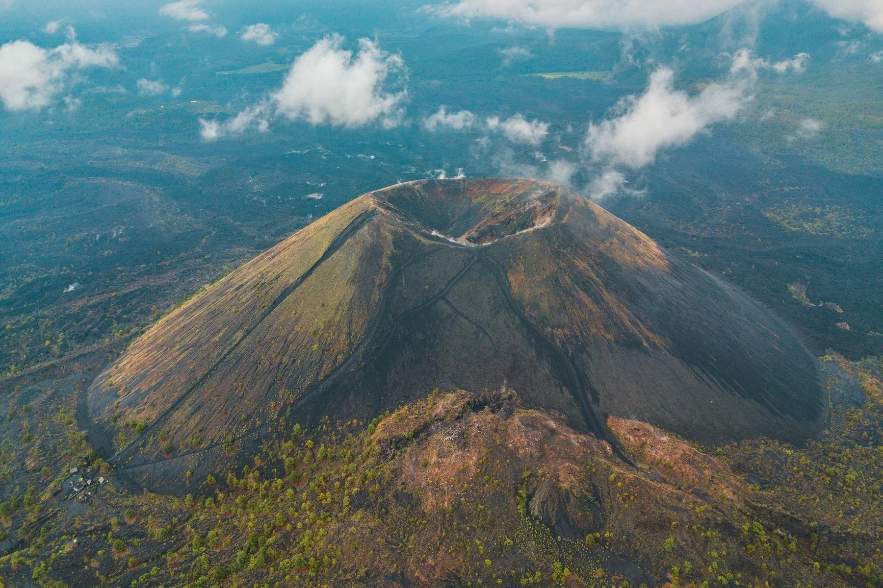 Photo aérienne du volcan Ibu à Malaisie, avec un cratère au sommet et des éruptions de vapeur ou de fumée. Le volcan est entouré de végétation dense et d'un paysage rural.