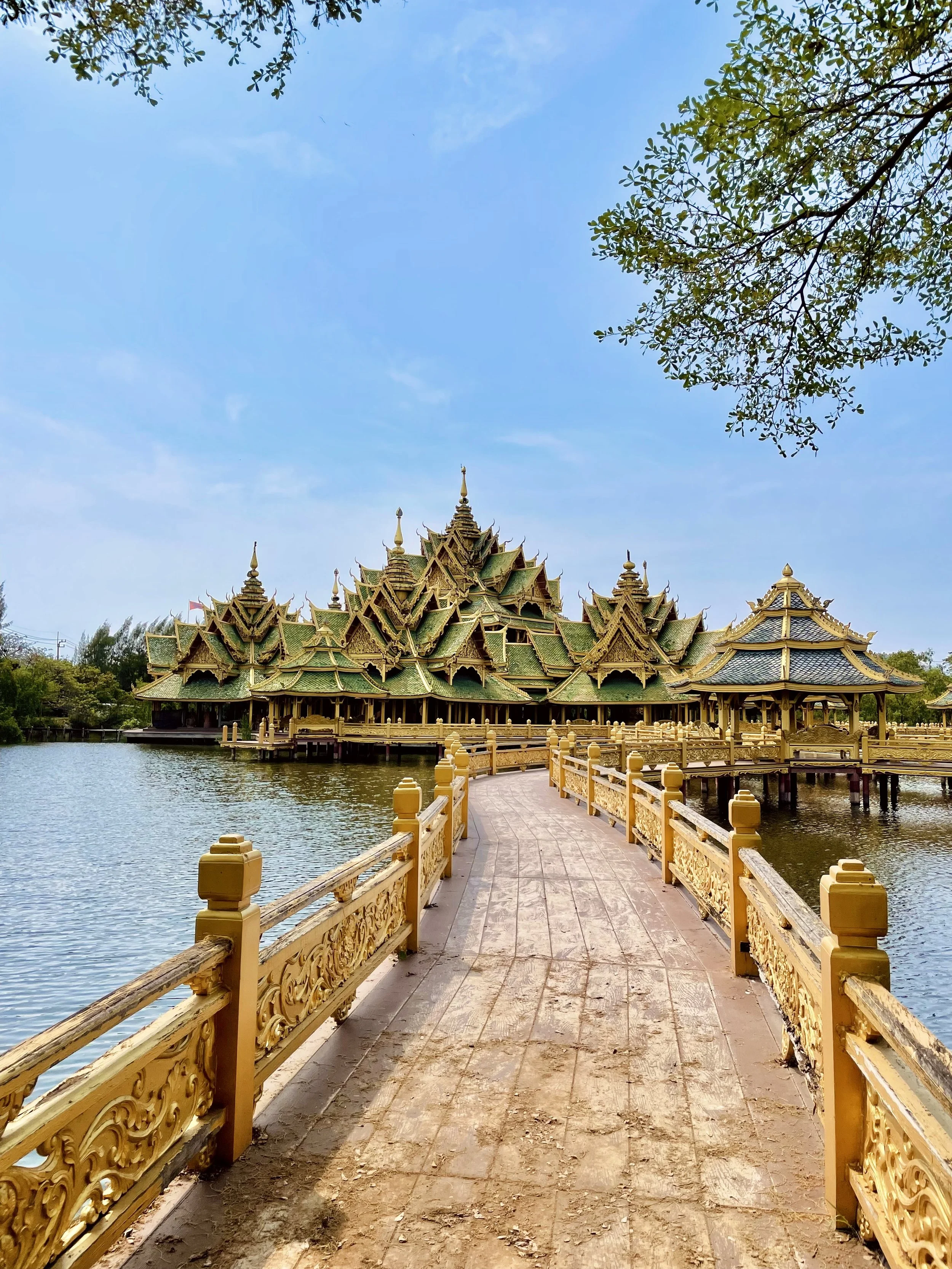 Un temple thaïlandais orné de détails dorés, situé sur l'eau, avec un pont en bois orné de sculptures en bois doré menant au temple, sous un ciel bleu.