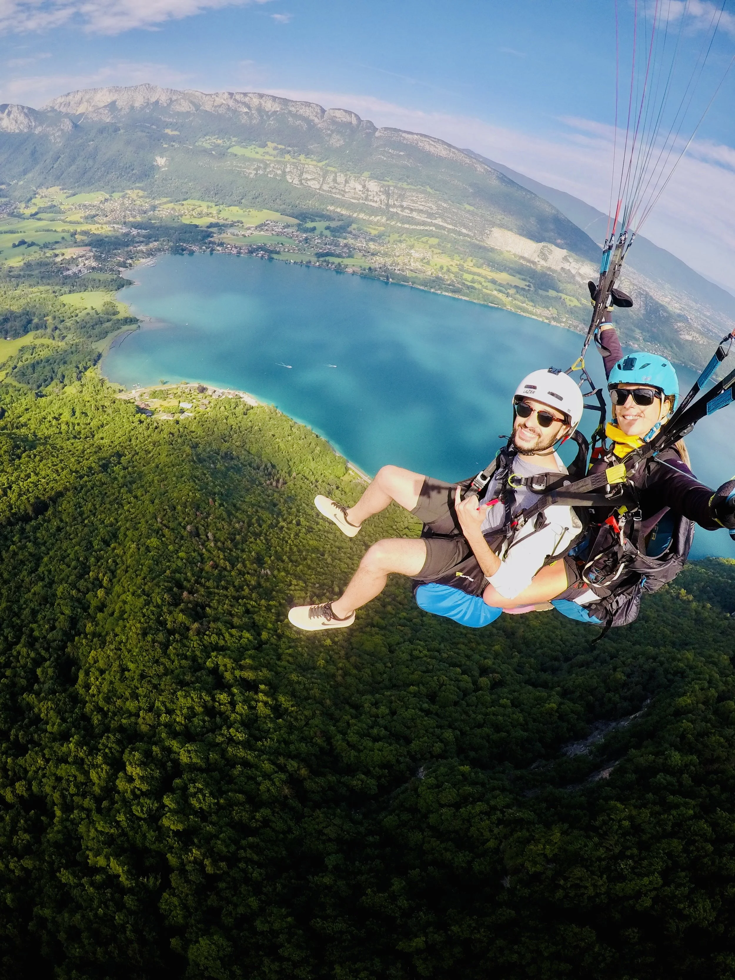 Deux personnes en tandem volant en parapente au-dessus d'une forêt verdoyante avec un lac bleu brillant et des montagnes en arrière-plan.