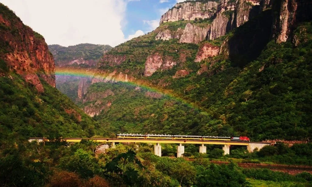 Un train traversant un pont au-dessus d'une vallée verdoyante, avec un arc-en-ciel enveloppant la montagne en arrière-plan.