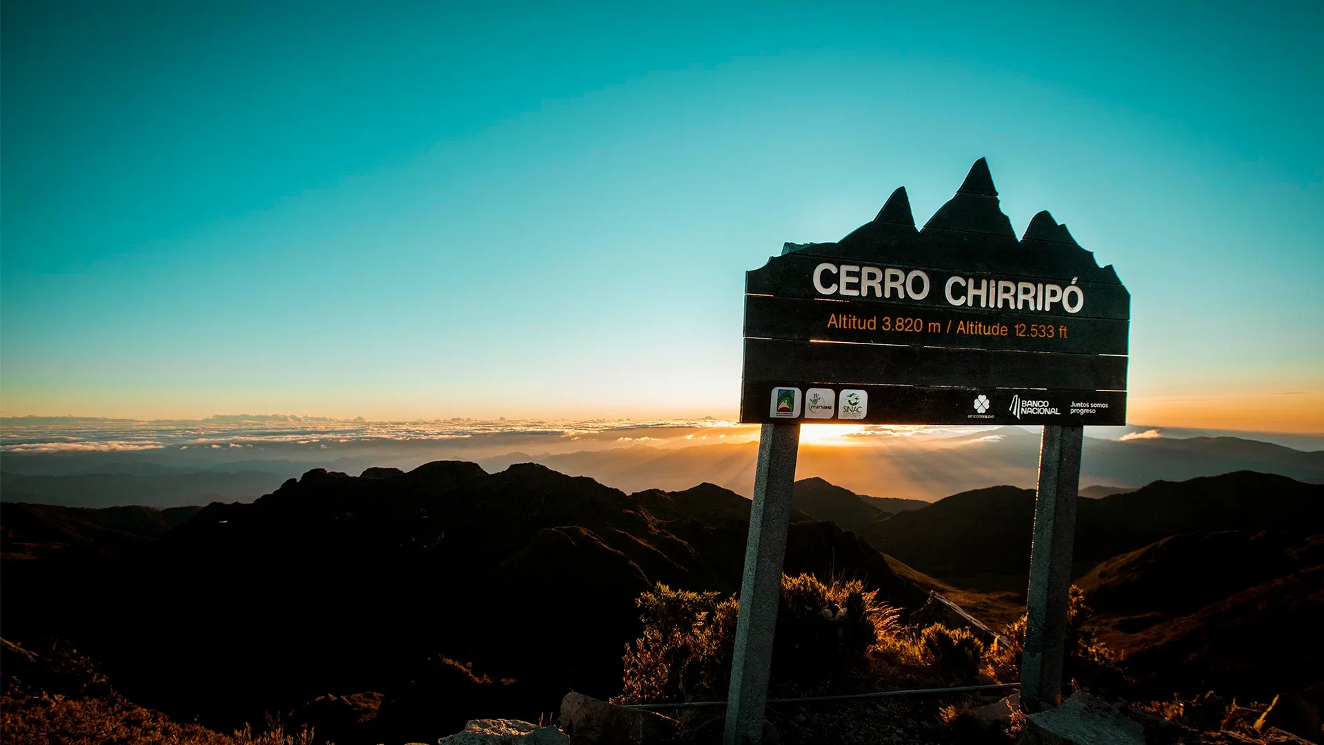 Panneau indiquant le sommet Cerro Chiripó à 3 820 mètres d'altitude, sur un fond de paysage montagneux au coucher du soleil avec des nuages et un ciel bleu clair.