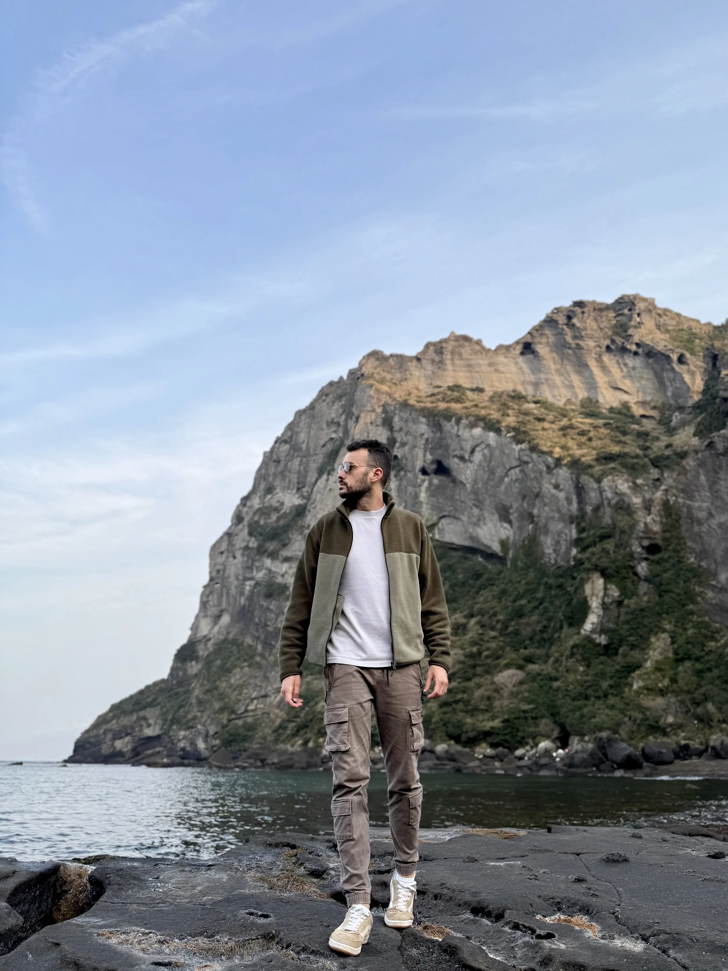 Un homme debout sur une plage rocheuse, portant une veste verte, un t-shirt blanc, un pantalon cargo gris, et des chaussures blanches, avec une montagne en arrière-plan, sous un ciel partiellement nuageux.