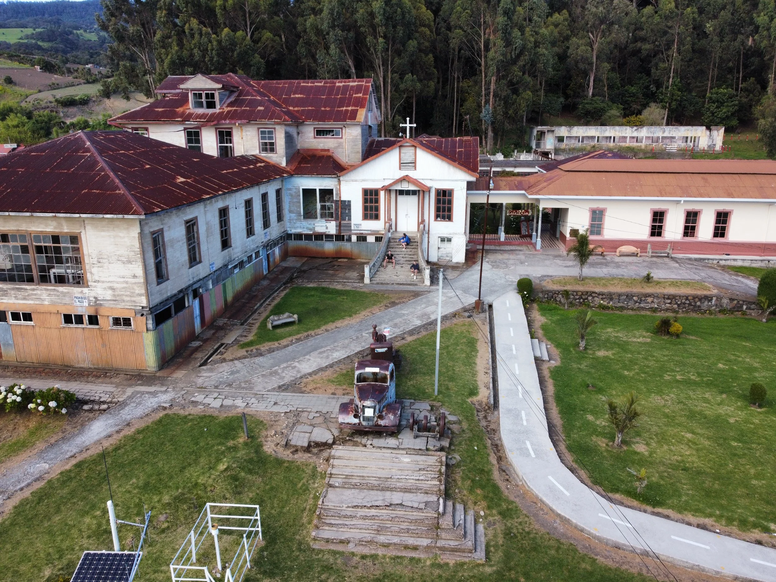 Une ancienne église en bois avec un toit rouillé et un bâtiment adjacent, entourée d’un espace vert avec des arbres, une route piétonne et un vieux camion stationné devant l’église.