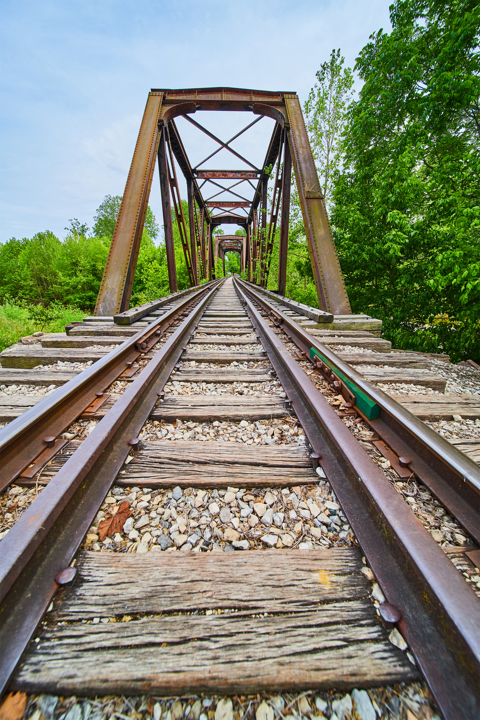 Old rusty railroad bridge with wooden planks and green trees on both sides, viewed from the ground level.