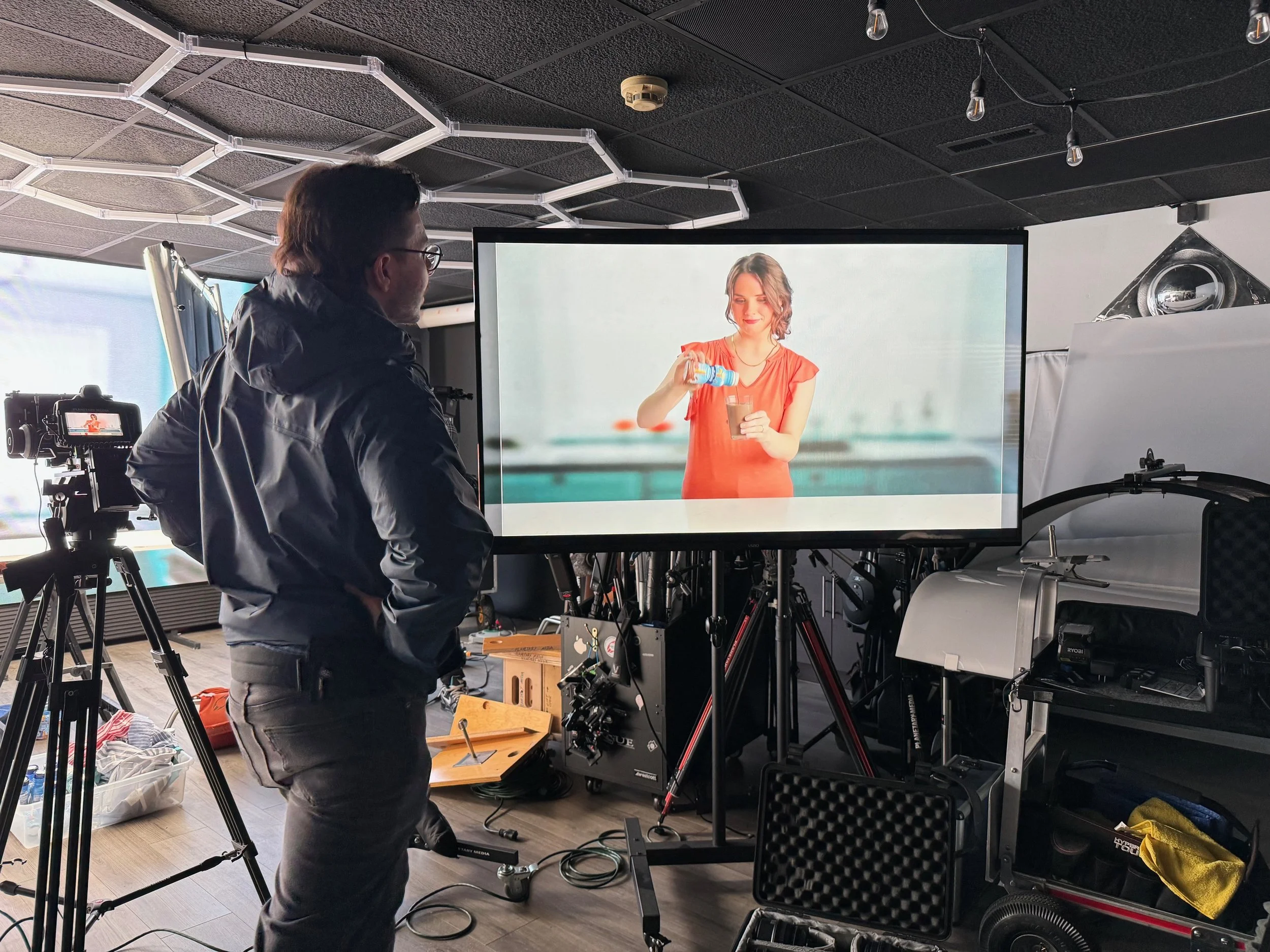 A man in a dark jacket and glasses stands in front of a camera and a large screen showing a woman pouring a drink, filming a video or photo shoot in a studio.