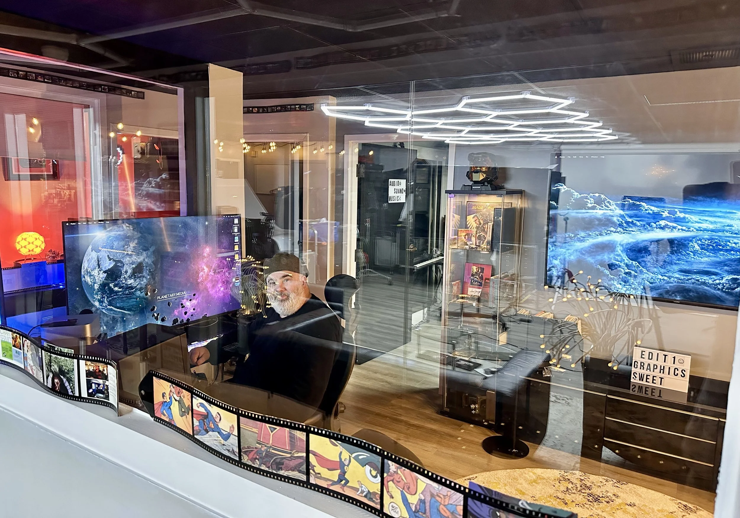 A man sits in a recording or editing studio, seen through a glass window. The studio contains multiple monitors displaying space-themed and cartoon images, with a large screen showing Earth from space. The room is decorated with modern lighting, book