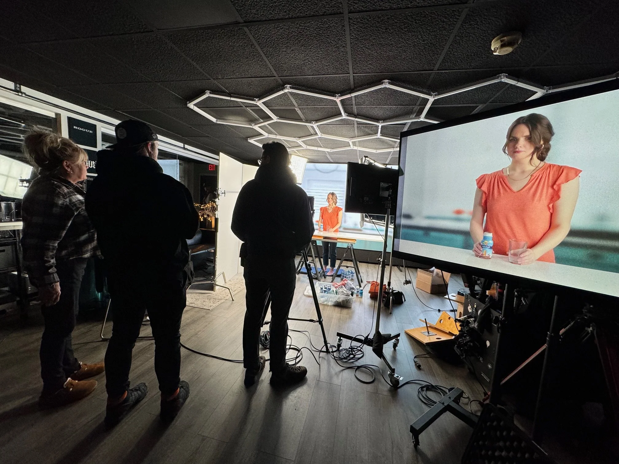 Behind-the-scenes of a video or photo shoot with three crew members watching a monitor showing a woman in an orange top holding a hydration bottle, setting up in a studio-like space with equipment and lighting.