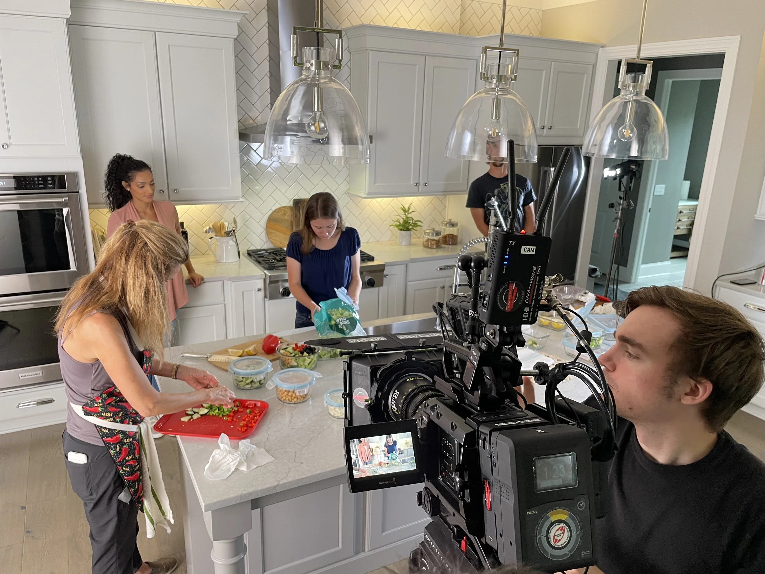 A woman in an apron chops vegetables at a kitchen island while three other women stand nearby; a man operates a professional video camera filming the scene, and studio lights illuminate the kitchen for a cooking video.