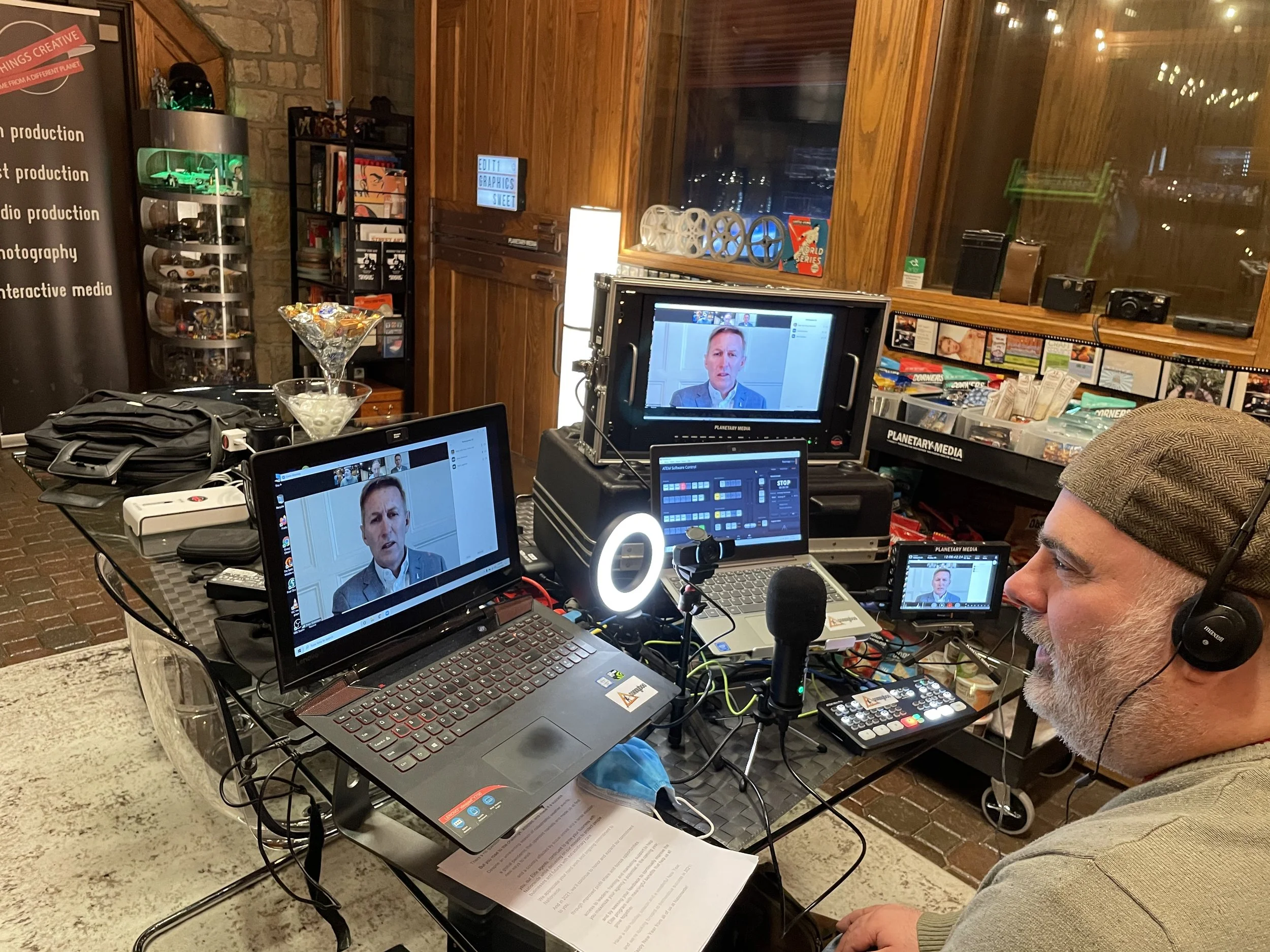 A man wearing headphones and a brown cap speaking in a video production studio with multiple monitors, cameras, and lighting equipment. The studio has wood paneling and a stone wall.