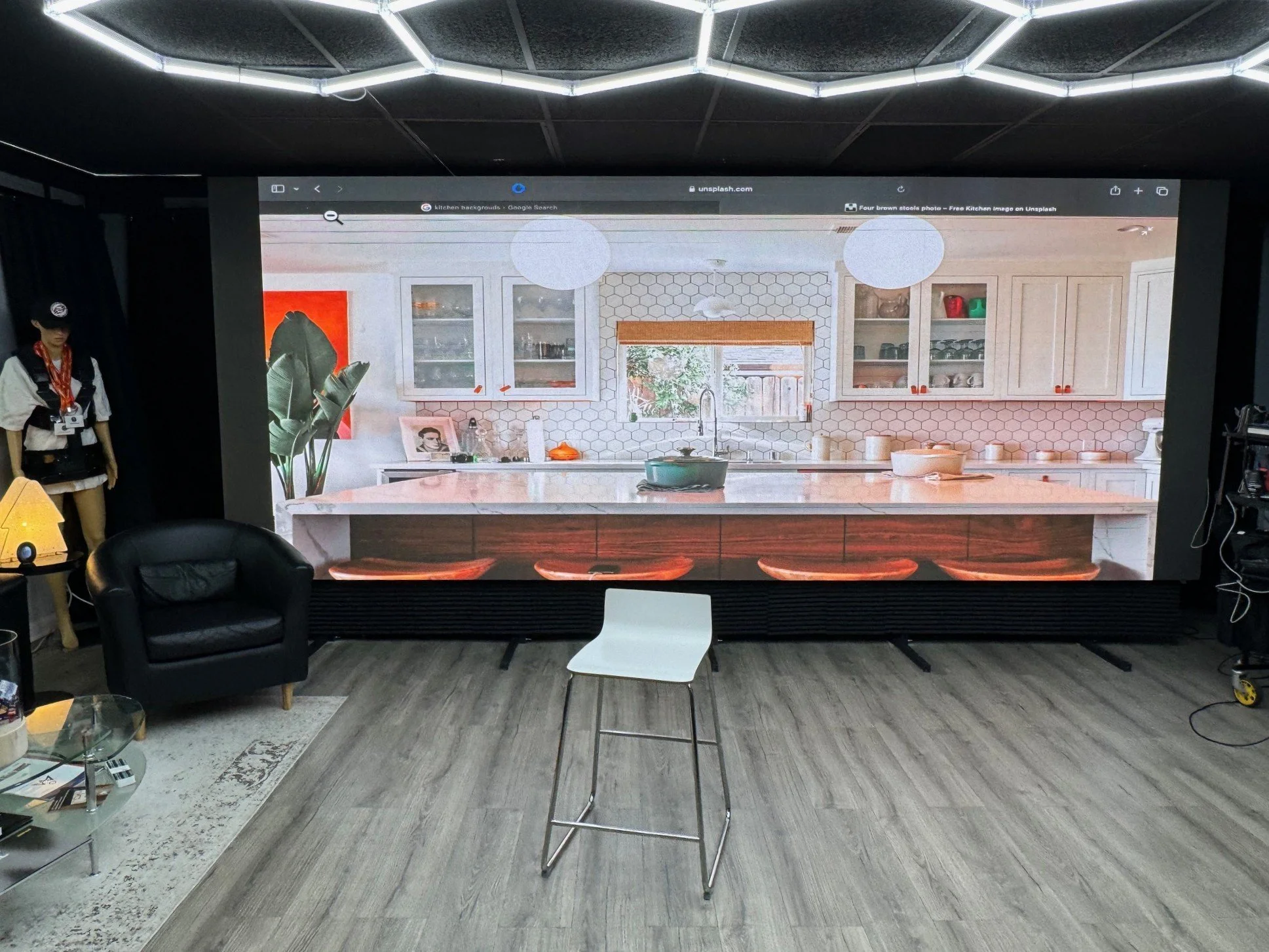 Large wall screen displaying a modern kitchen with white cabinets, a marble island, and a honeycomb tile backsplash. The real room has a gray wood floor, a black armchair, a white chair, and nearby furniture.
