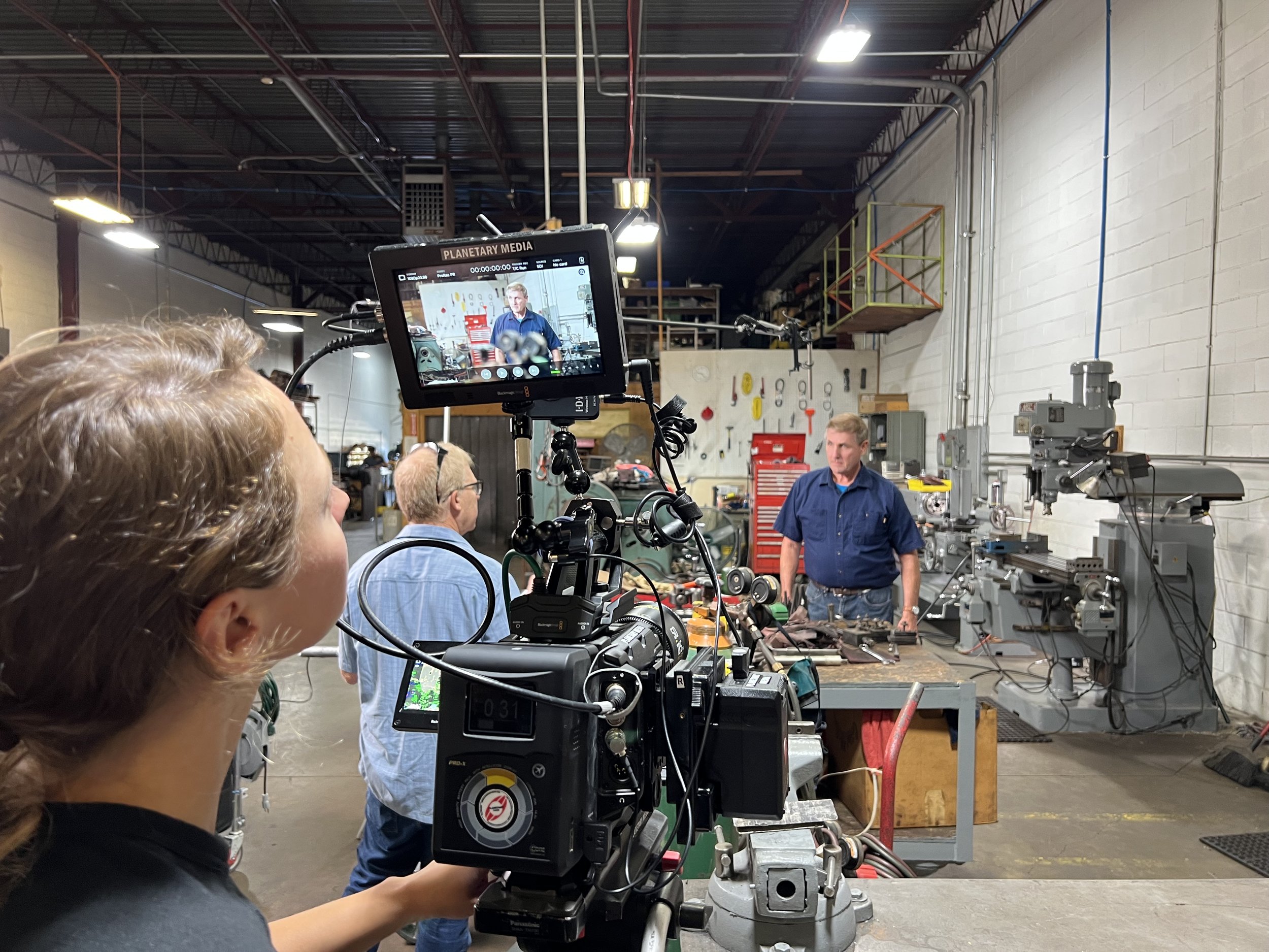 A video crew filming a man in a blue shirt in a workshop with various tools and machinery, with the camera's monitor showing the scene being recorded.