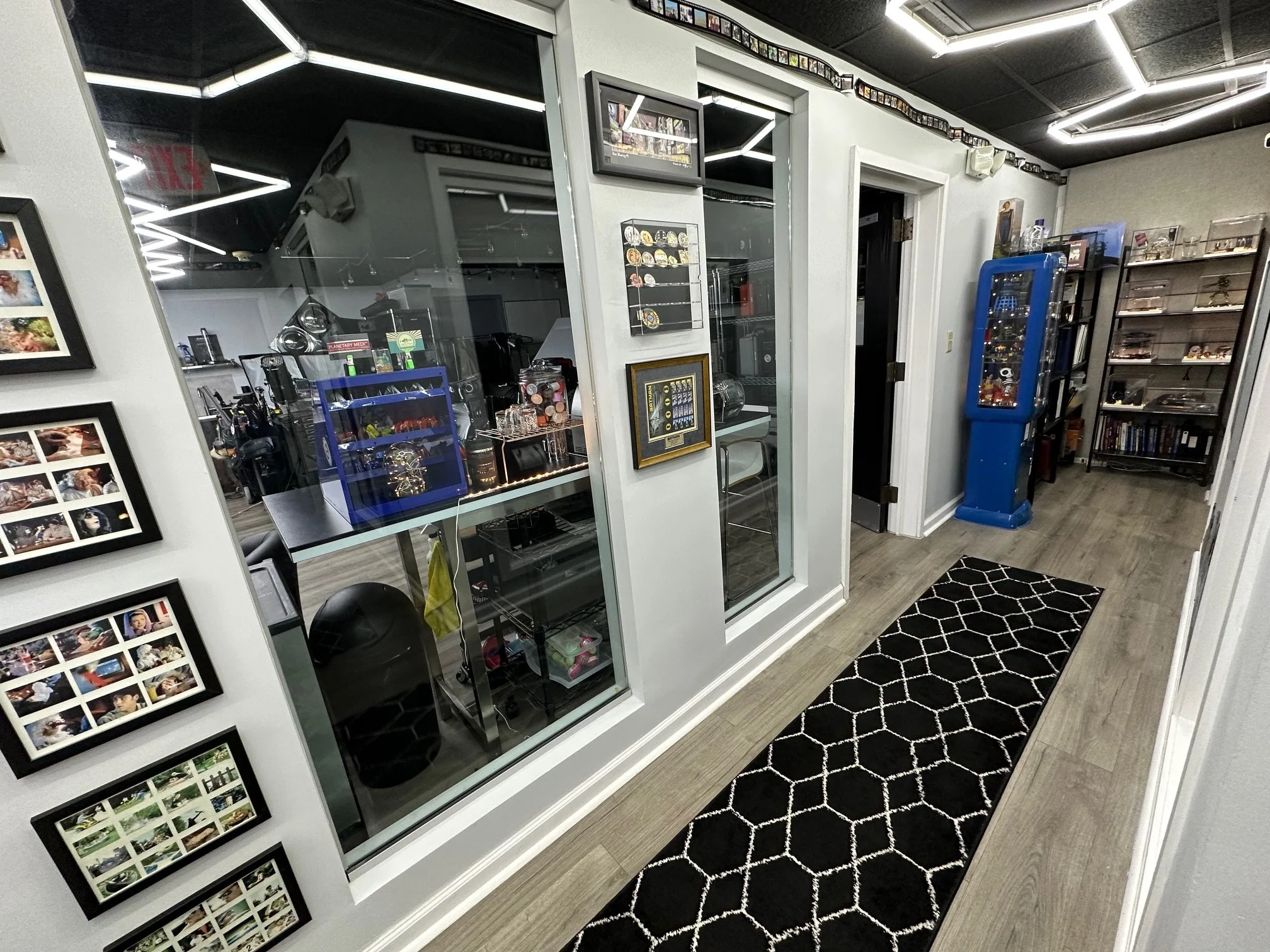 View of an interior room with a hallway, black and white patterned rug, and display shelves filled with collectibles and books. Behind a glass wall, a room with various items including photo frames and a blue display case. Wall decorations include fr