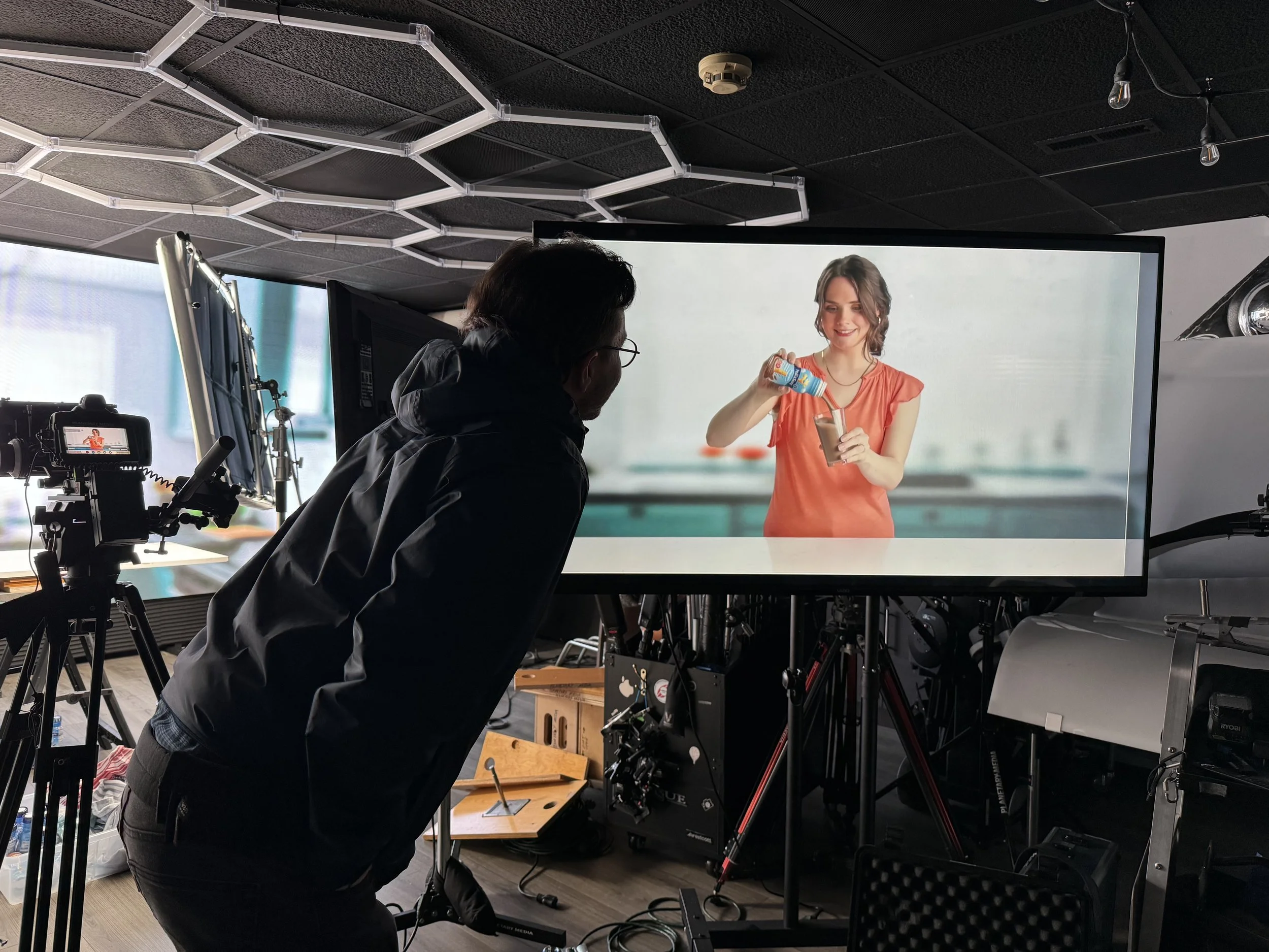 A man on a film set watching a woman in a kitchen pouring a beverage into a glass from a bottle on a large screen.