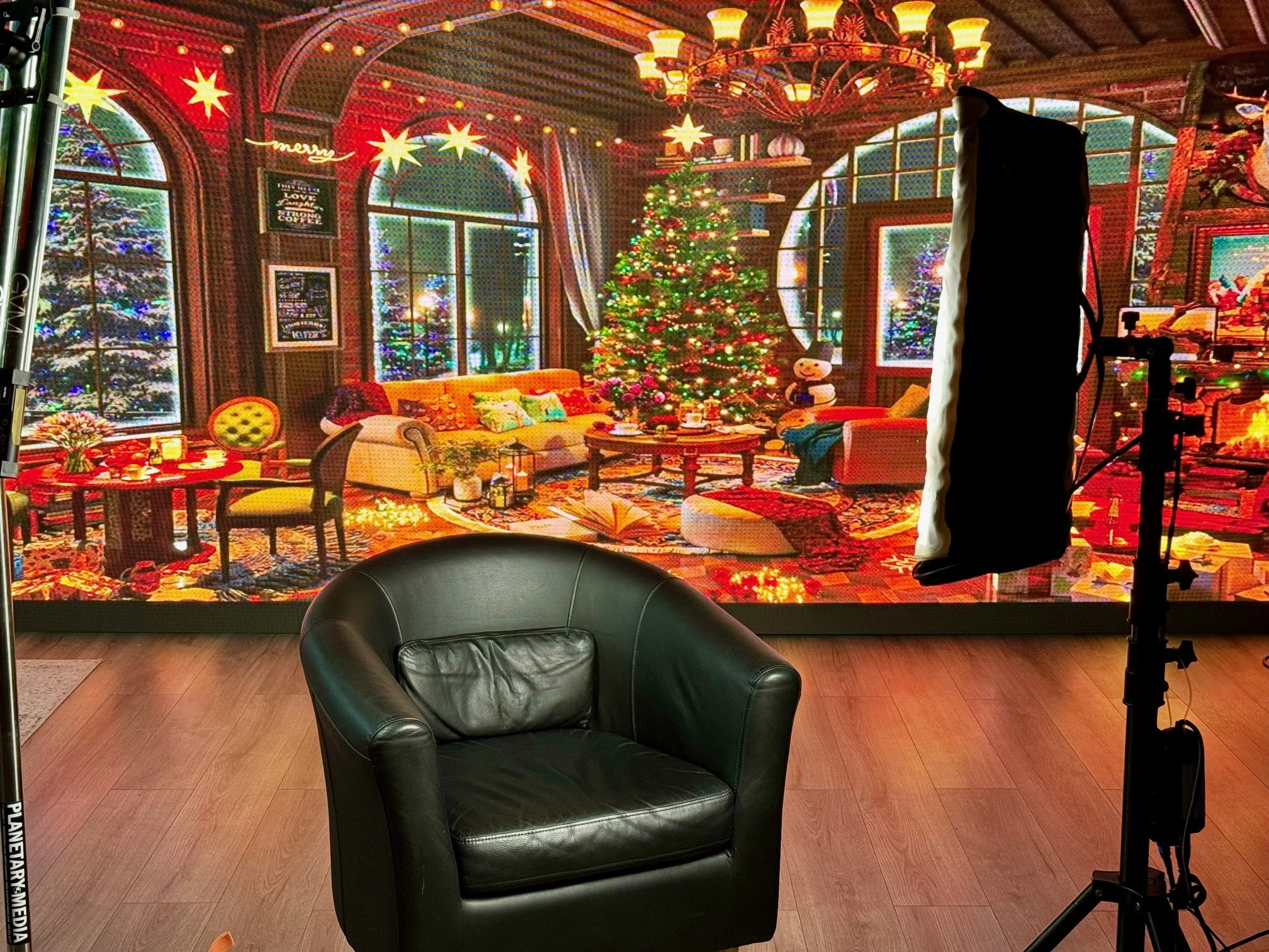 A cozy living room decorated for Christmas with a lit Christmas tree, colorful ornaments, wrapped presents, a snowman figure, and festive string lights. Large windows show a snowy outdoor scene.
