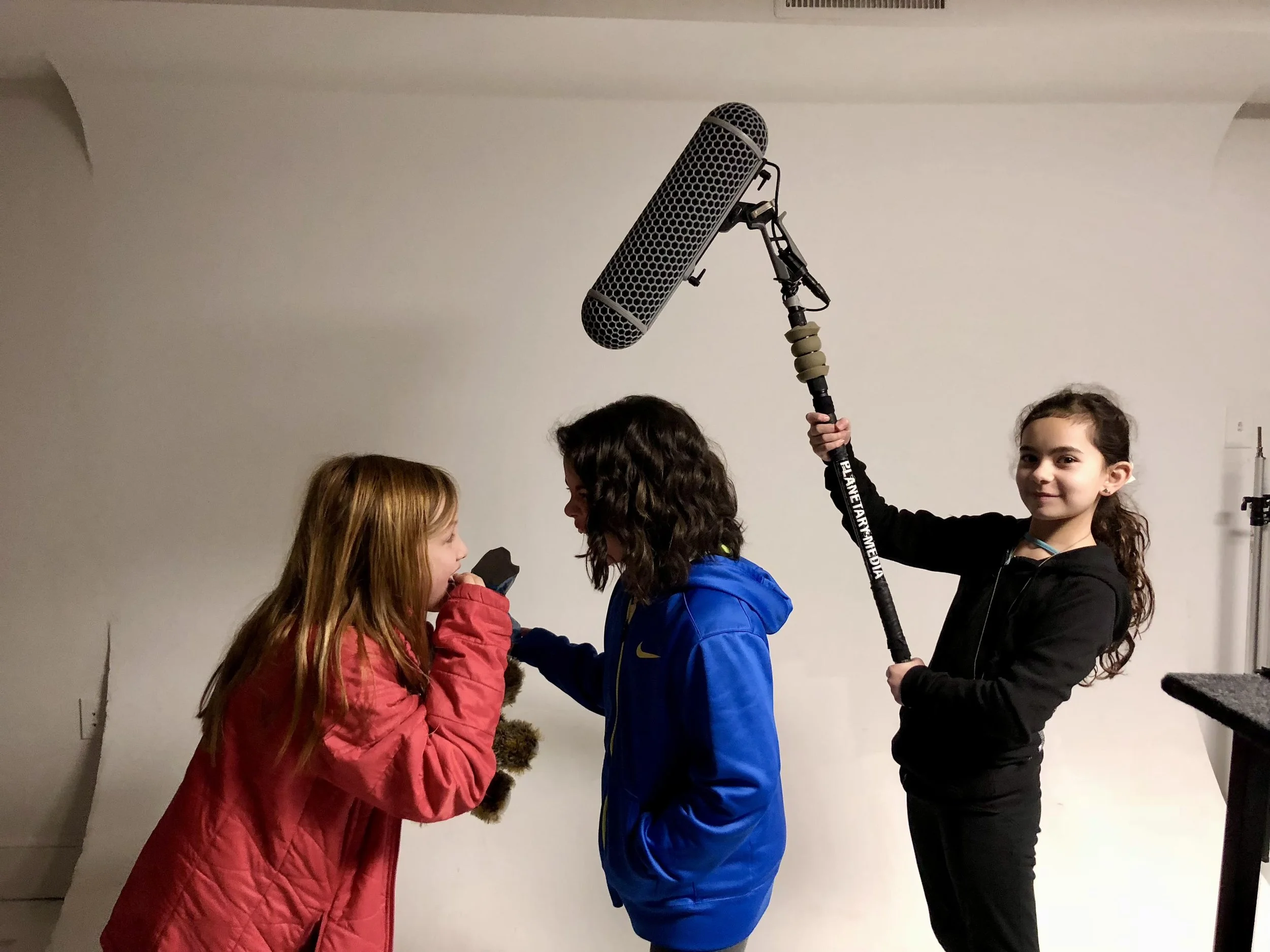 Three girls in a recording studio, one holding a boom microphone over the other two, who are facing each other and speaking into a microphone.