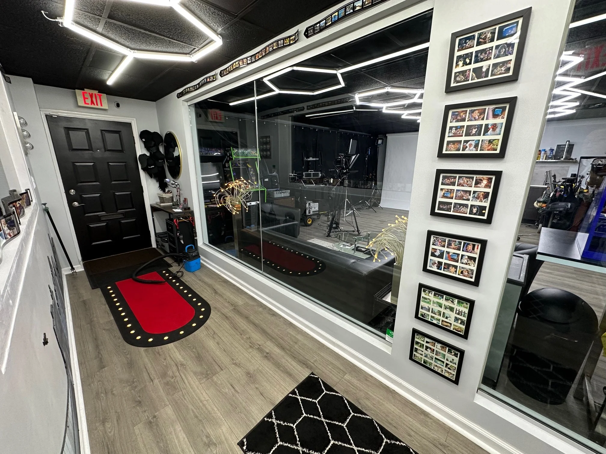 View of a modern fitness or photo studio with a black door, wall-mounted equipment, framed photo collages, hardwood floors, and a glass wall overlooking a recording or workout area with fitness equipment and cameras.