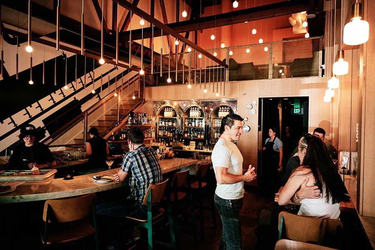 People socializing at a bar inside a modern restaurant with wood decor, hanging lights, and a backdrop of shelves with bottles.