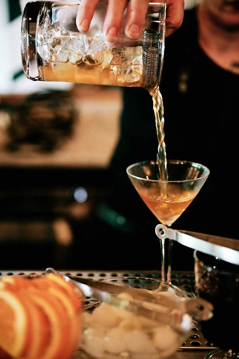 A bartender pouring a cocktail from a mixing glass with ice into a martini glass. In the foreground, there is an orange and a small bowl of ice cubes. The background is slightly blurred.