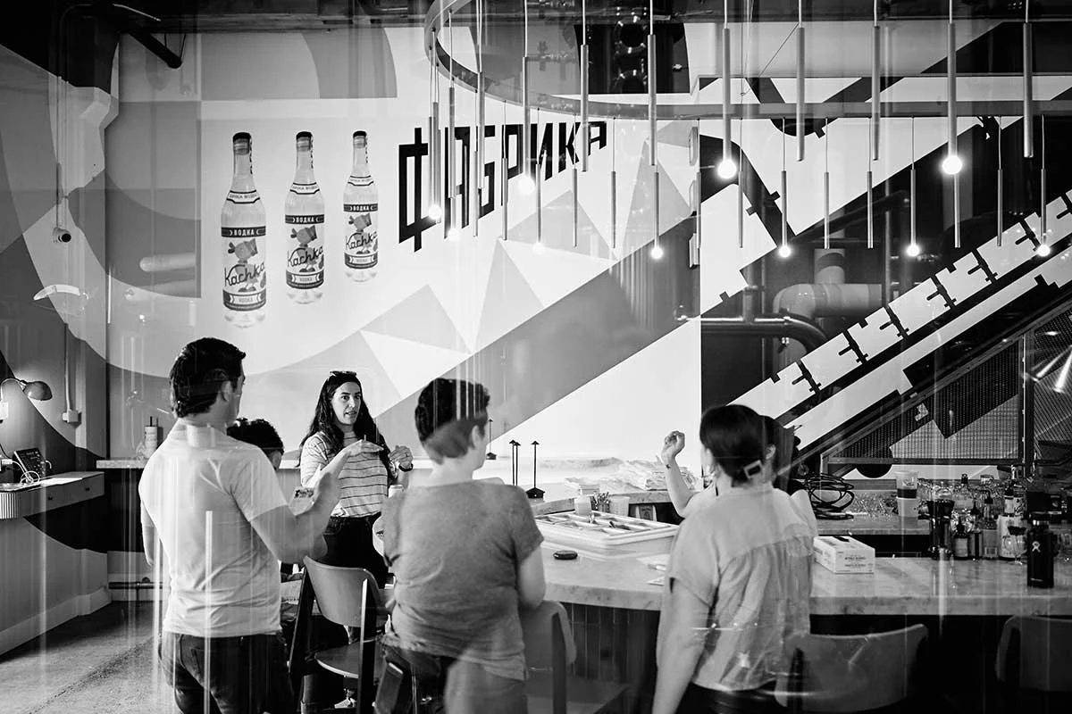 A group of people having a discussion in a modern, industrial-style cafe or bar, with a mural on the wall depicting bottles and Russian text.
