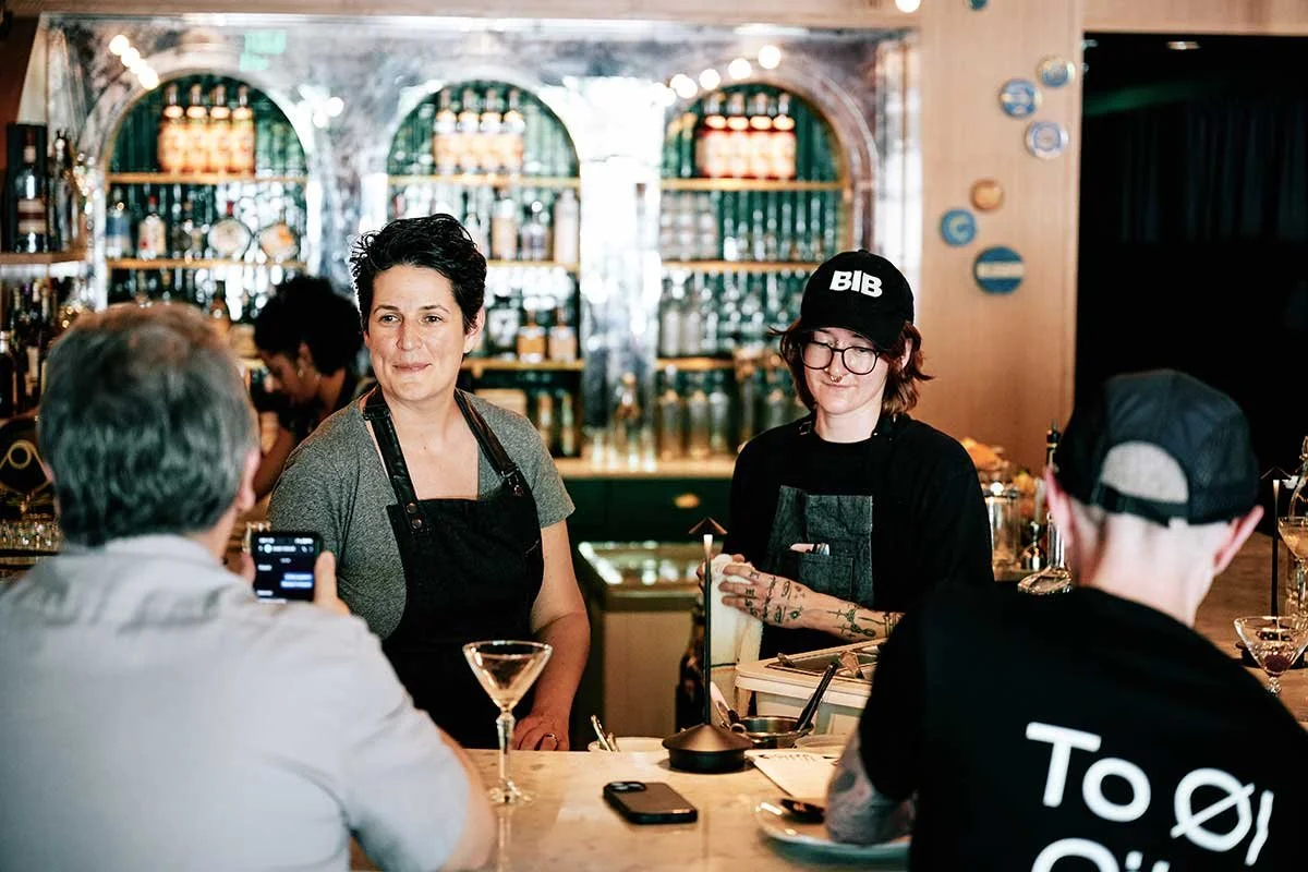 A woman barista with short hair and a gray shirt, and a young female with glasses, tattoos, and a black cap that says 'BIB', are working behind the bar in a restaurant. They are serving drinks to customers who are seated at the bar, while a couple of customers use their phones. The background shows shelves with bottles and glasses.