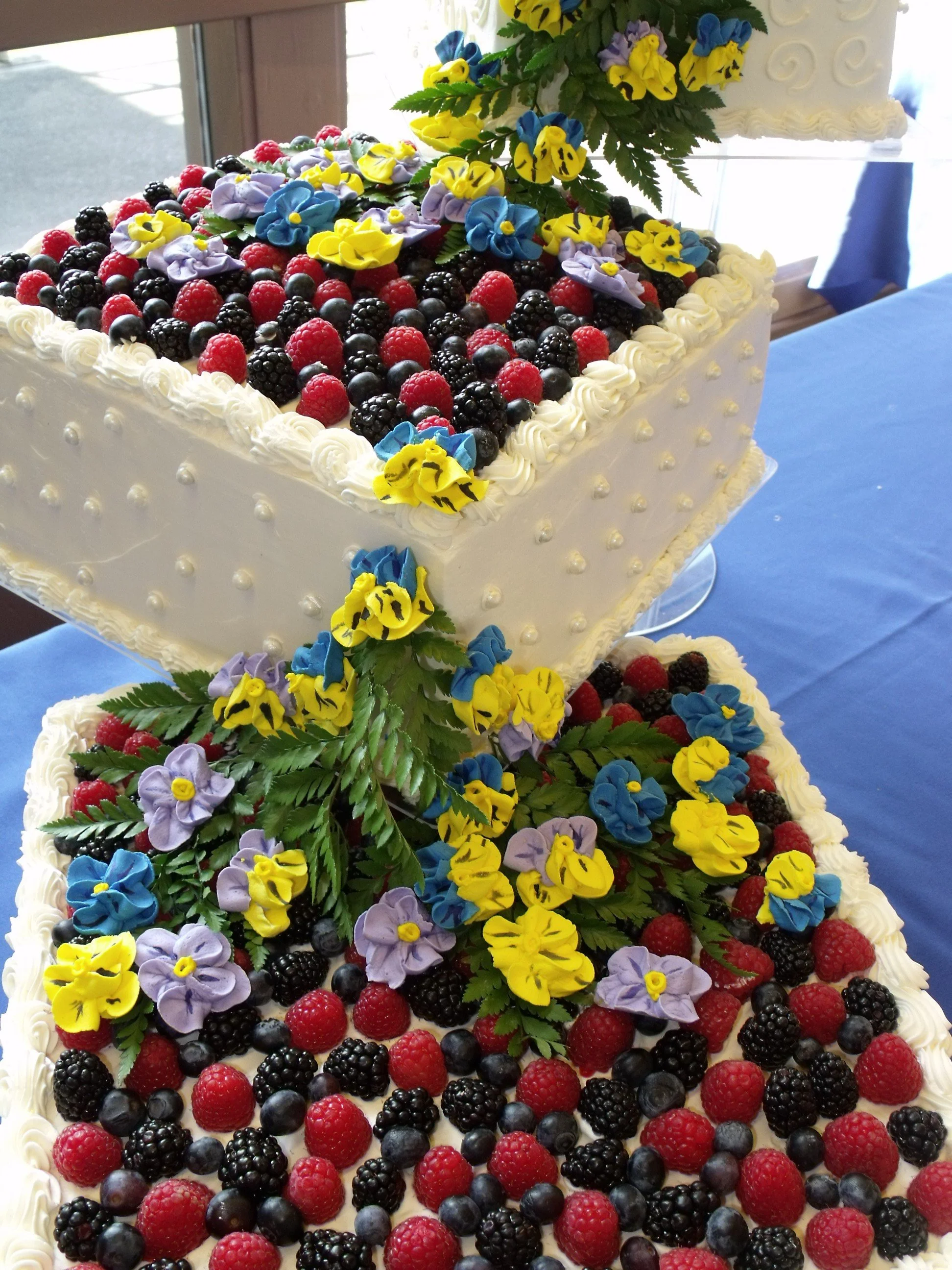 Three Tier Wedding Cake Fresh Berries Atop Each Of Three Crystal Pillar Separated Square Buttercream Cake Tiers Piped Dot and Scroll Side Decor Flowing Sugar Porcelain Pansy Cascade