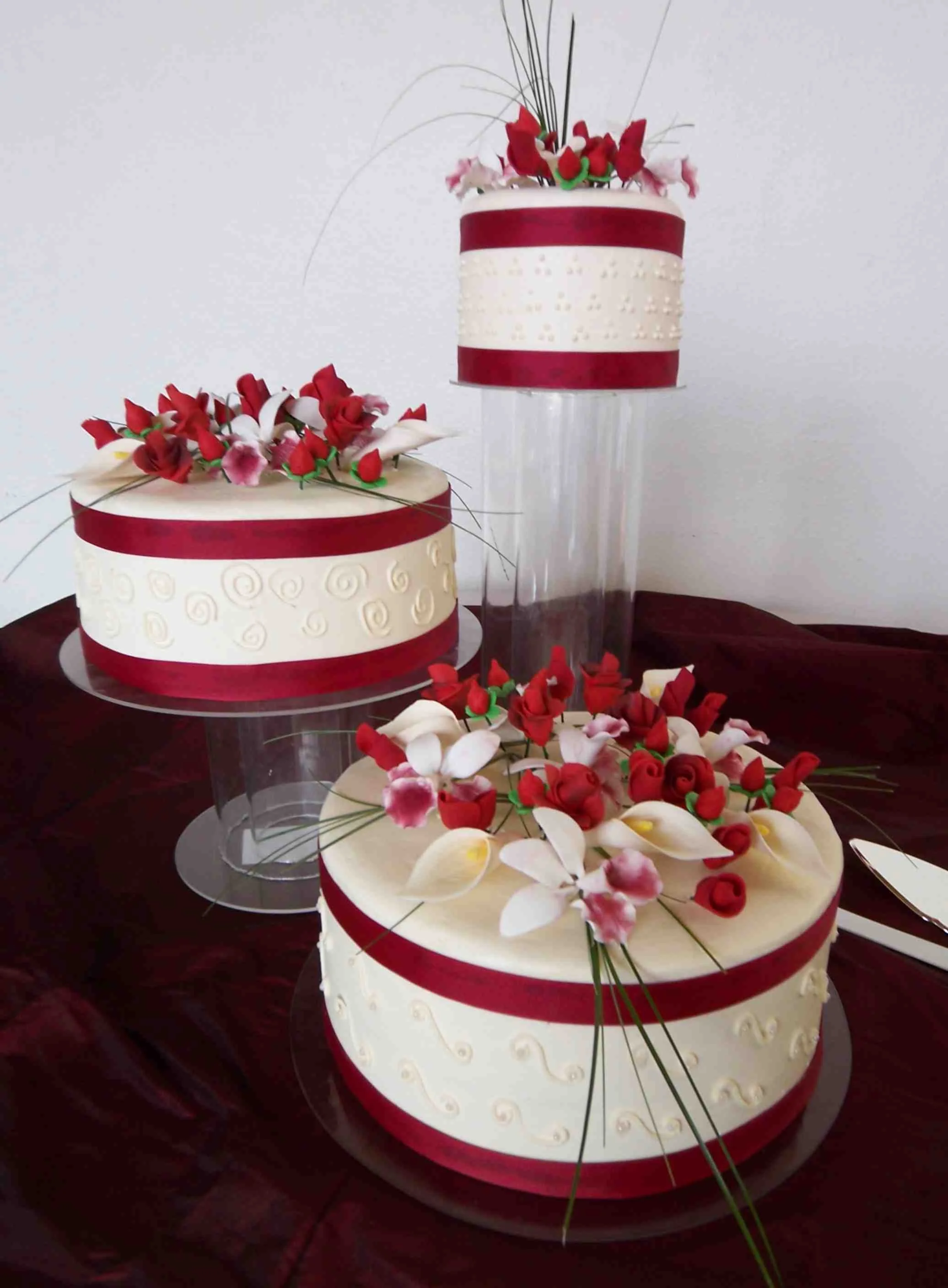 Three independent round buttercream frosted wedding cakes crystal cake plates risers red satin ribbons top and bottom of cake sides piped scrolls pom arrangements topping each of cakes sugar porcelain red bud roses small white Callas white Stargazer 