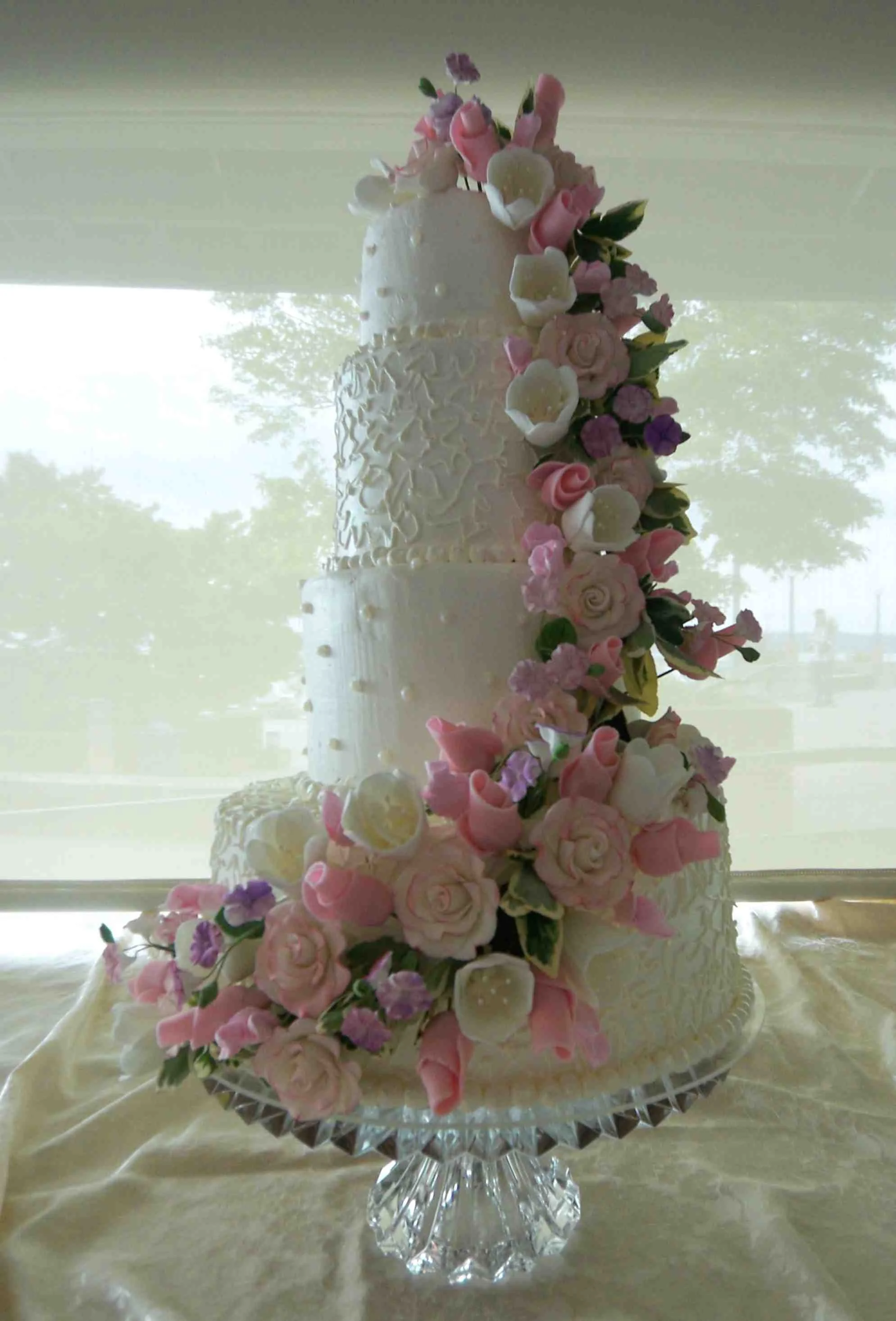 Four Tier stacked round tiers wedding cake on glass cake pedestal abundant flowing sugar porcelain floral arrangement from top to bottom sugar dusted pink bud and full open roses white tulips hydrangea blossoms  stephanotis over variegated fresh ivy 