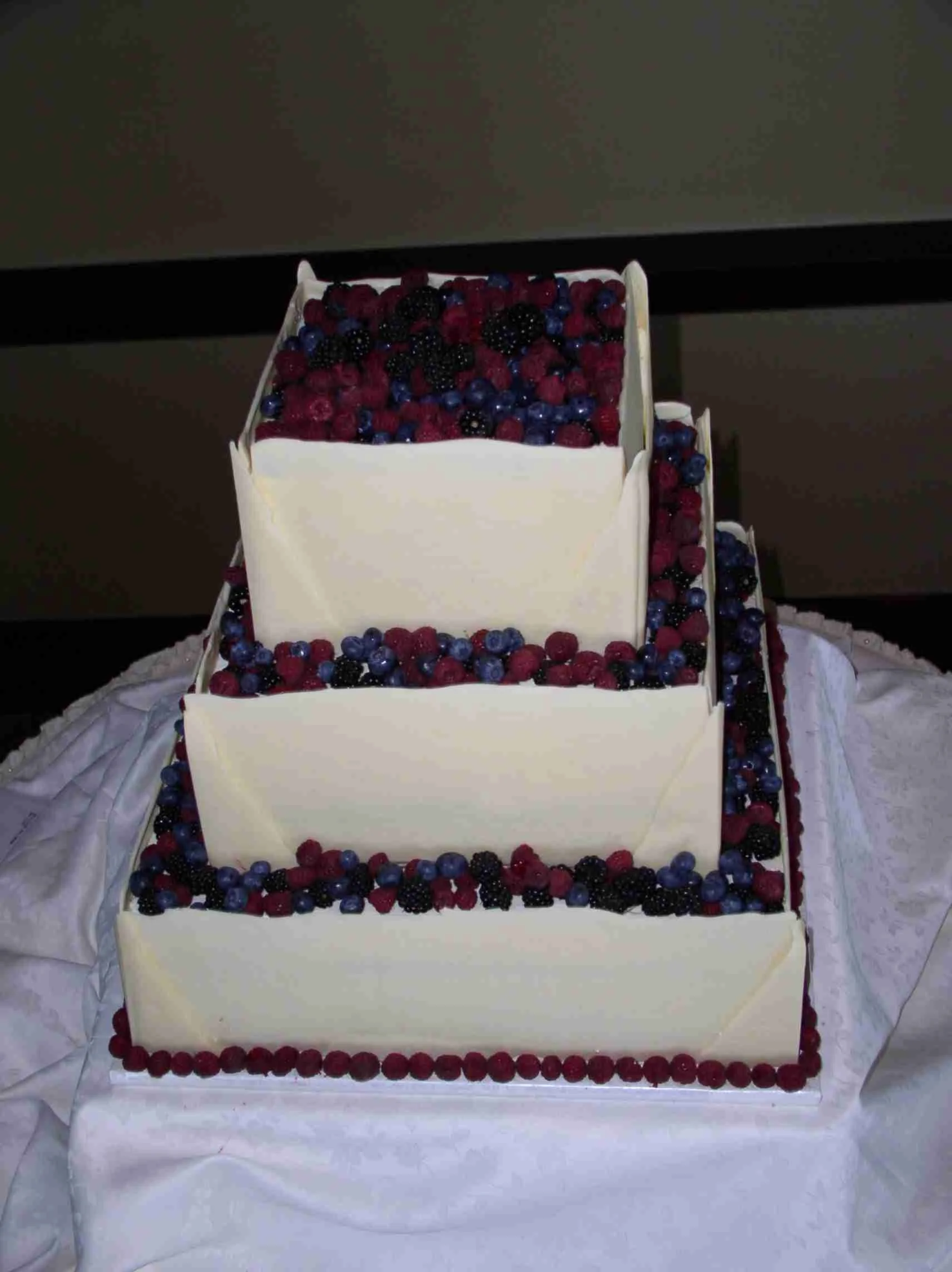 Wedding Cake Fresh Berries Atop Three Square Stacked Cake Tiers with White Chocolate Sheets on Sides and Corners to Hold Blue Berries Raspberries with Raspberry Bottom Border