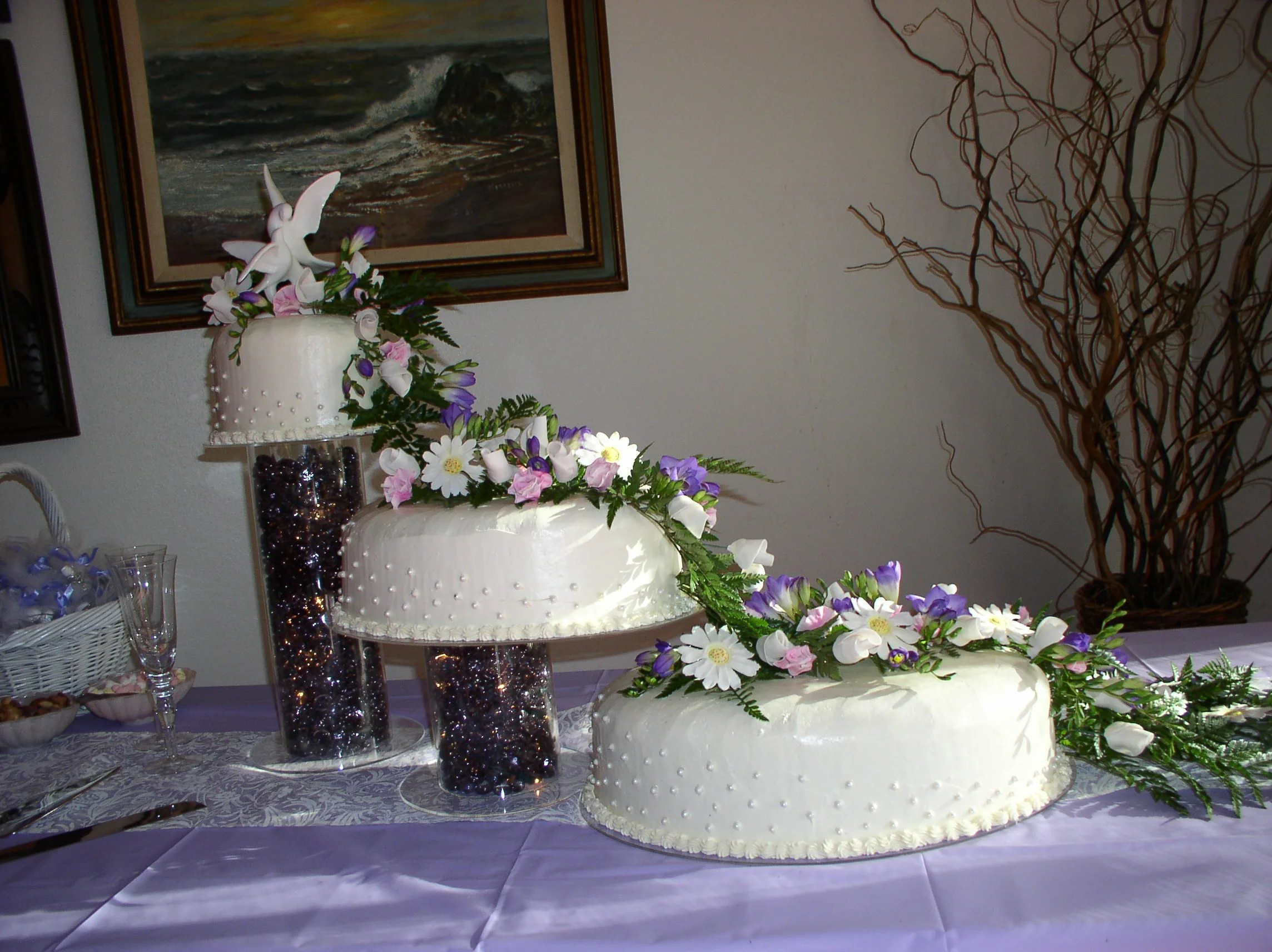 Three independent oval buttercream rounded top edge wedding cake crystal plate and risers colored stone filled columns flowing fresh greens sugar dusted sugar porcelain pink roses daisies crocus clear piped dots diagonal placement sides of cake  pipe