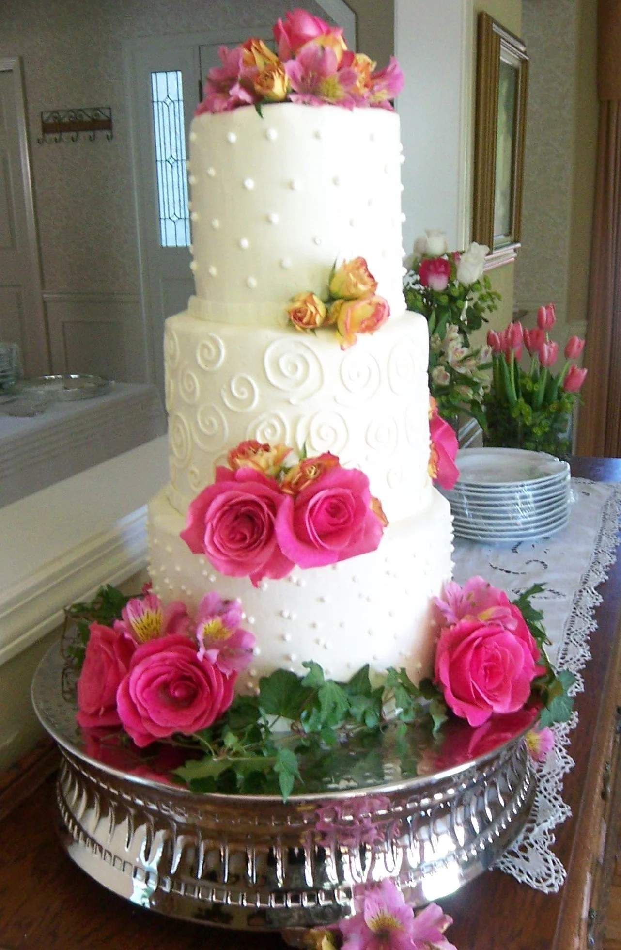 Elegant Wedding Cake Three Tier Stacked Round Smooth Buttercream Frosted Tiers with No Borders Pipped Side Designs of Scrolls and Pearl Dots Cake is Sitting on Silver Cake Pedestal Green Ivy Vines at Base of Cake and Pom Placements of Pink Full Open 