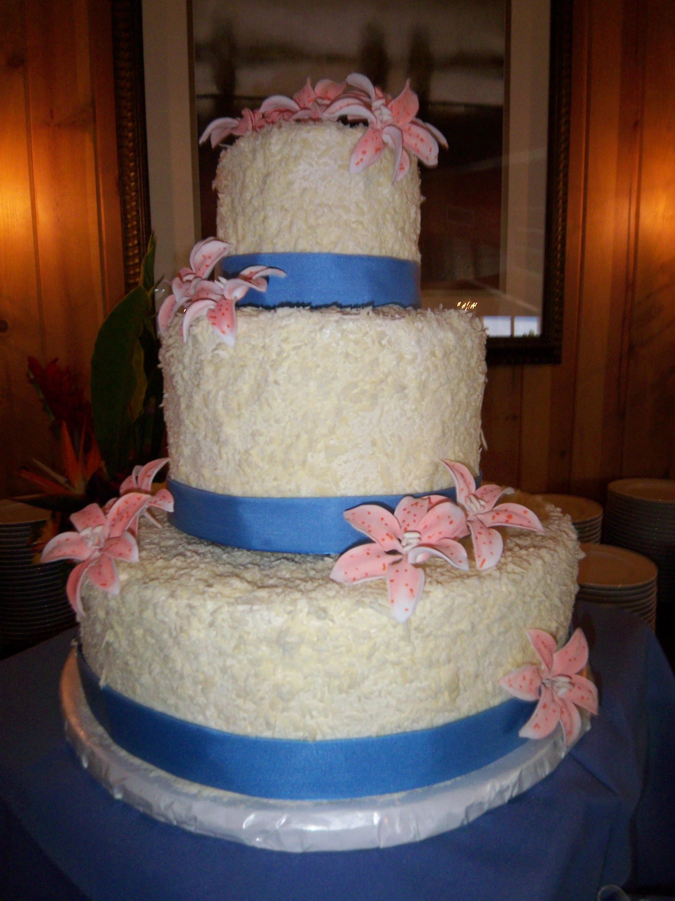 Three tier wedding cake round buttercream frosted stacked round cakes tops and sides encrusted with coconut Blue satin ribbons surround bottom of cakes simple placement of pink sugar dusted sugar porcelain Stargazer Lilly's